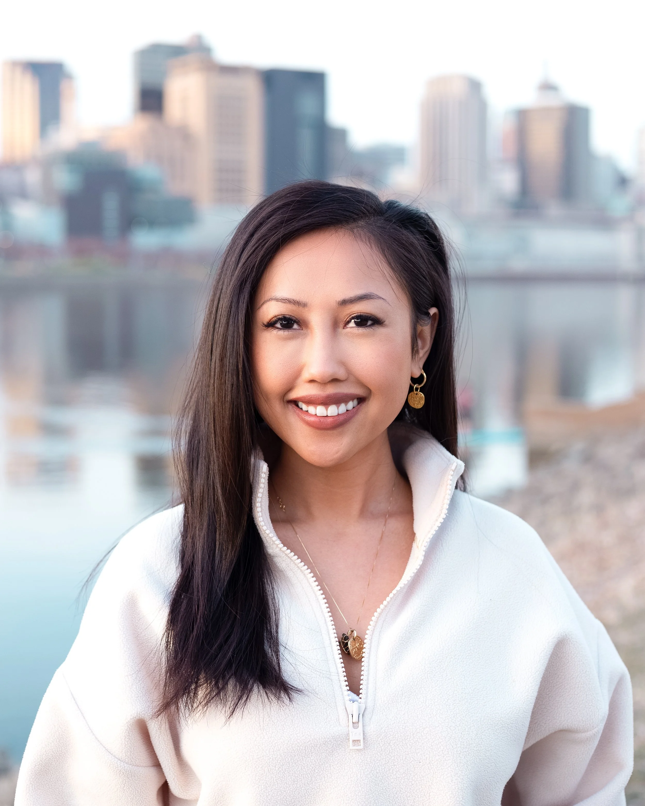 Ammada Tuy smiling outdoors with the downtown Saint Paul skyline and river in the background.