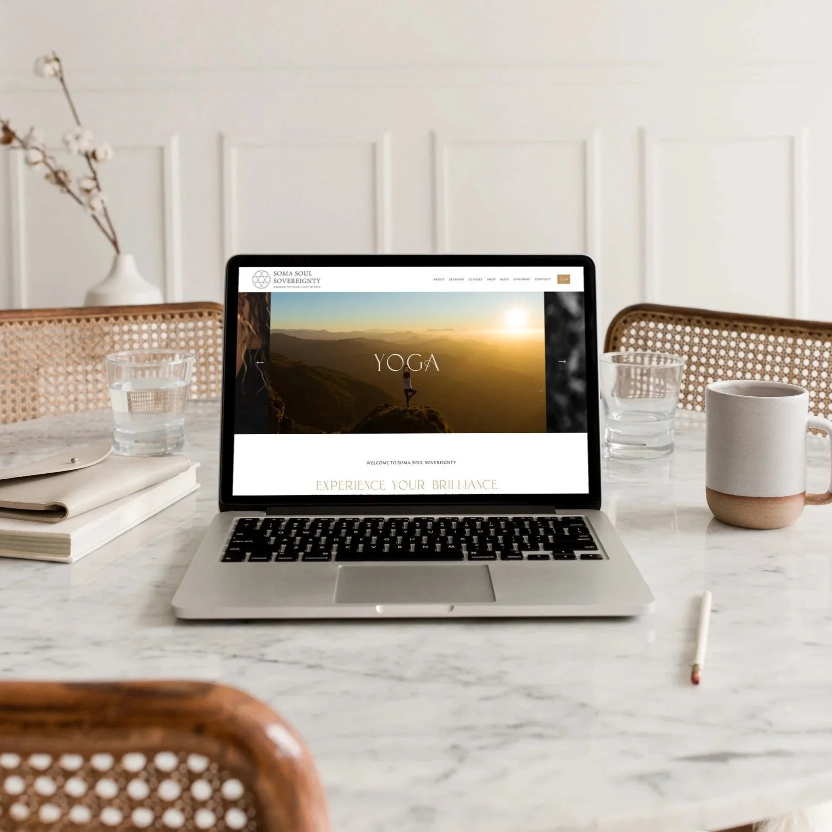 A laptop on a marble dining table displaying a yoga website, with notebooks, glasses of water, a ceramic mug, and a pen around it.