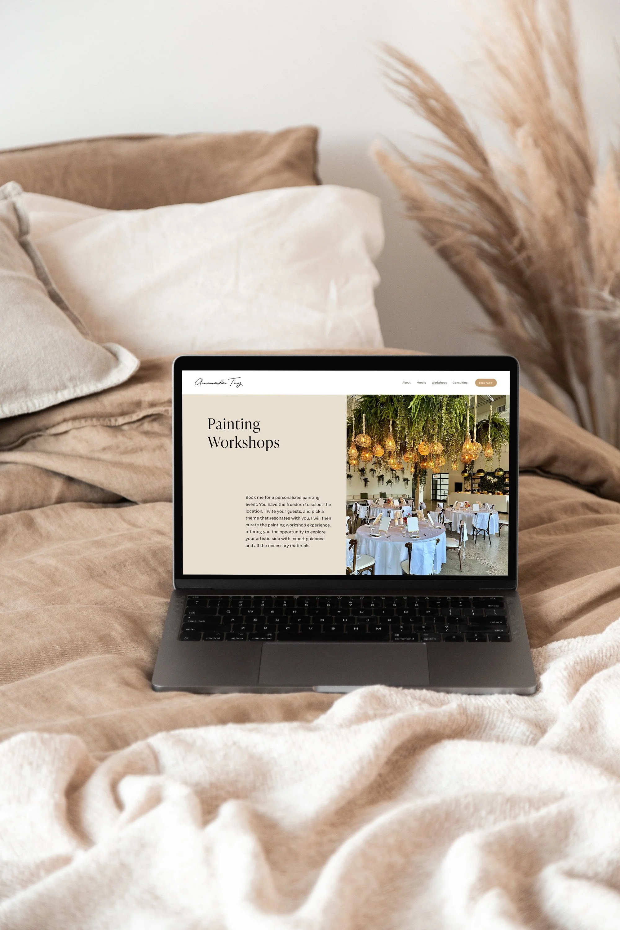 A laptop on a beige bedspread displaying a website for painting workshops, with a decorated dining area visible on the screen.