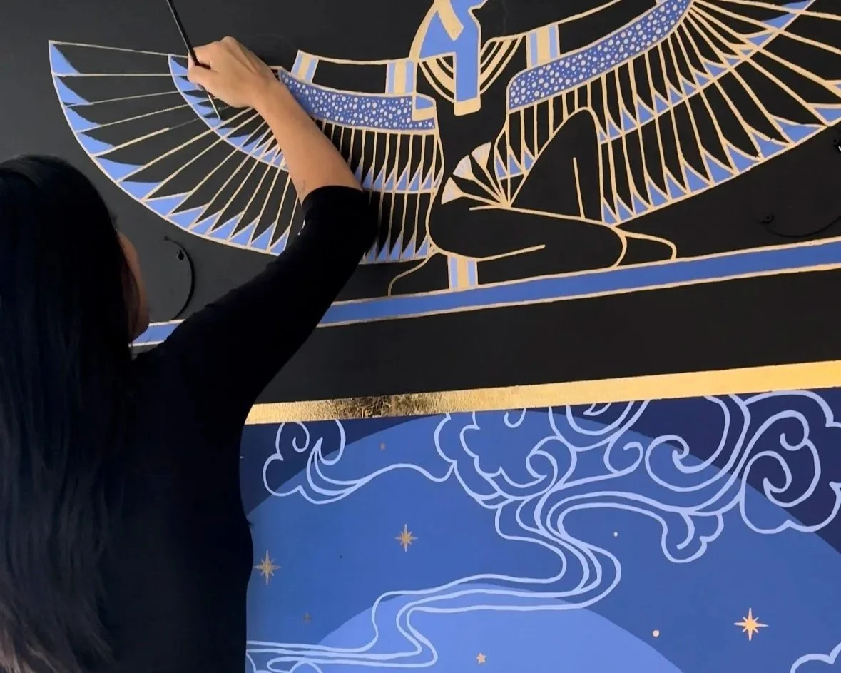 Person painting a mural featuring a stylized Egyptin goddess with outstretched wings, geometric patterns, and celestial elements.