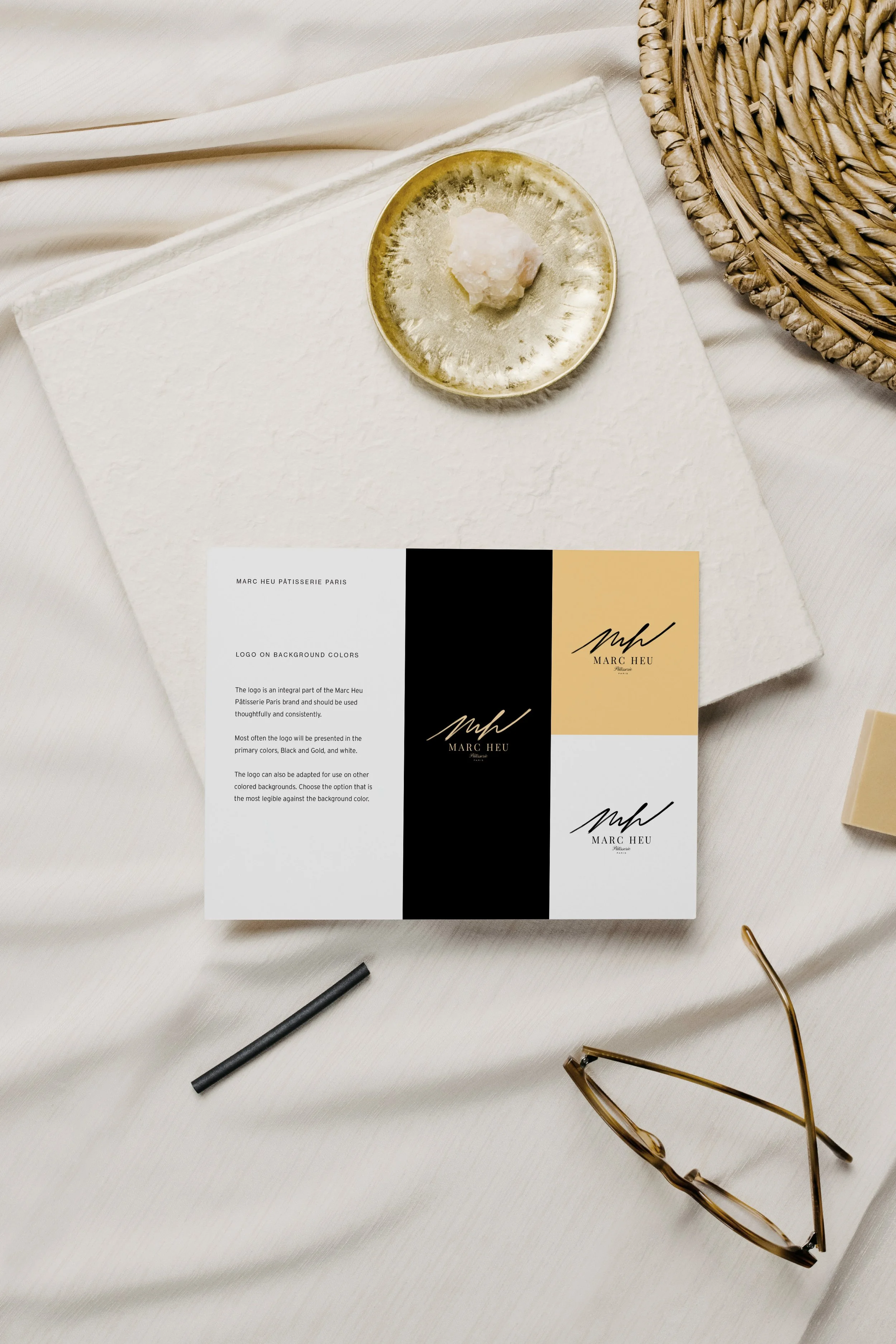 A flat lay image shows a white textured surface with a gold dish holding a pinkish-white substance, a piece of white cloth, a black pen, a pair of tortoiseshell glasses, and a printed branding guide for Marc Heu Patisserie Paris with black, gold, and white color blocks.