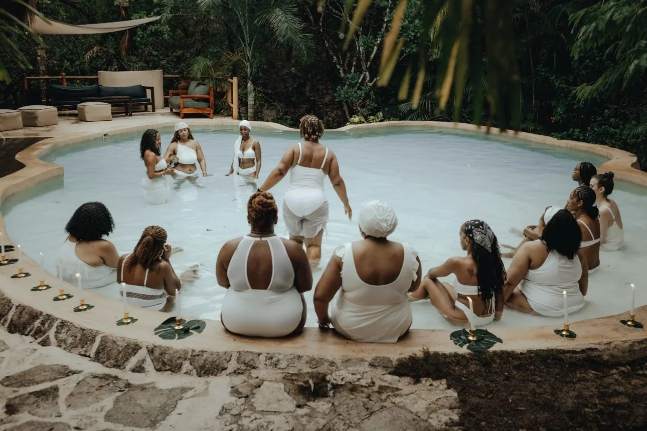 Women seated in circle around the edge of a milky pool during a water ceremony, candles before them, Ashley in the water at the center.