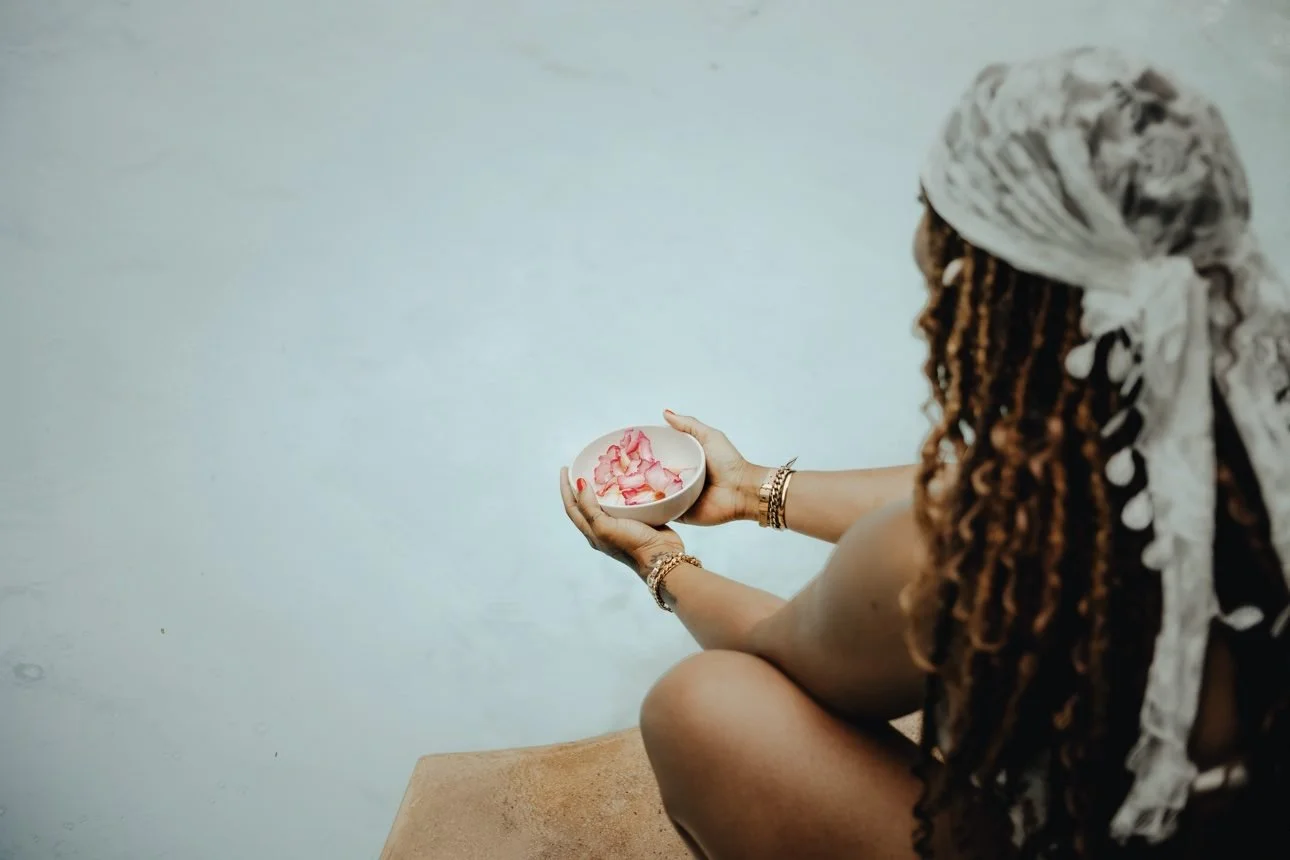 Seen from behind, Ashley cradles a small white bowl of pink rose petals above milky water, her honey locs and white lace headwrap trailing — her photograph.