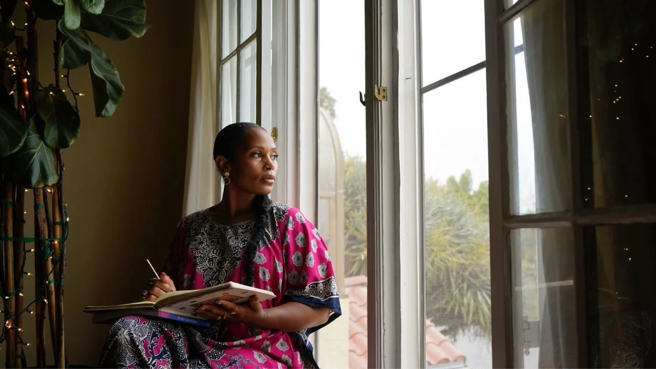 Ashley seated by tall windows in a magenta patterned dress, journal open and pen in hand, gazing out — her photograph.