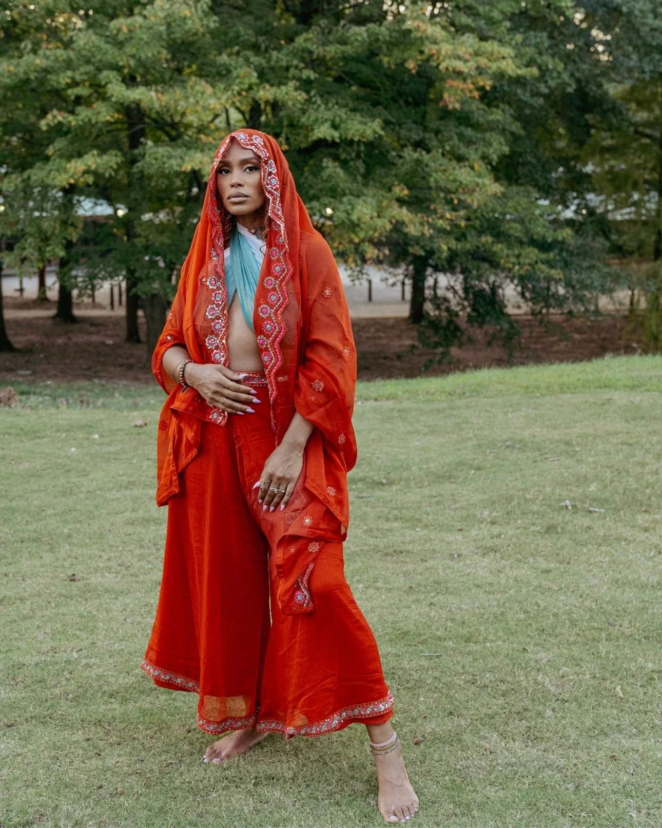 Ashley Curtis standing barefoot in the grass in a burnt-orange embroidered outfit, an orange veil over her head and one hand resting on her womb, trees behind her — her own photograph.