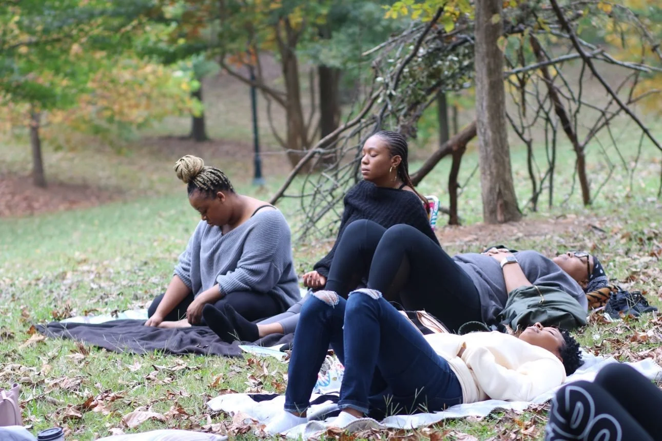 Participants resting on blankets beneath autumn trees while Ashley sits upright at the center in black, eyes closed, holding the space — her photograph.