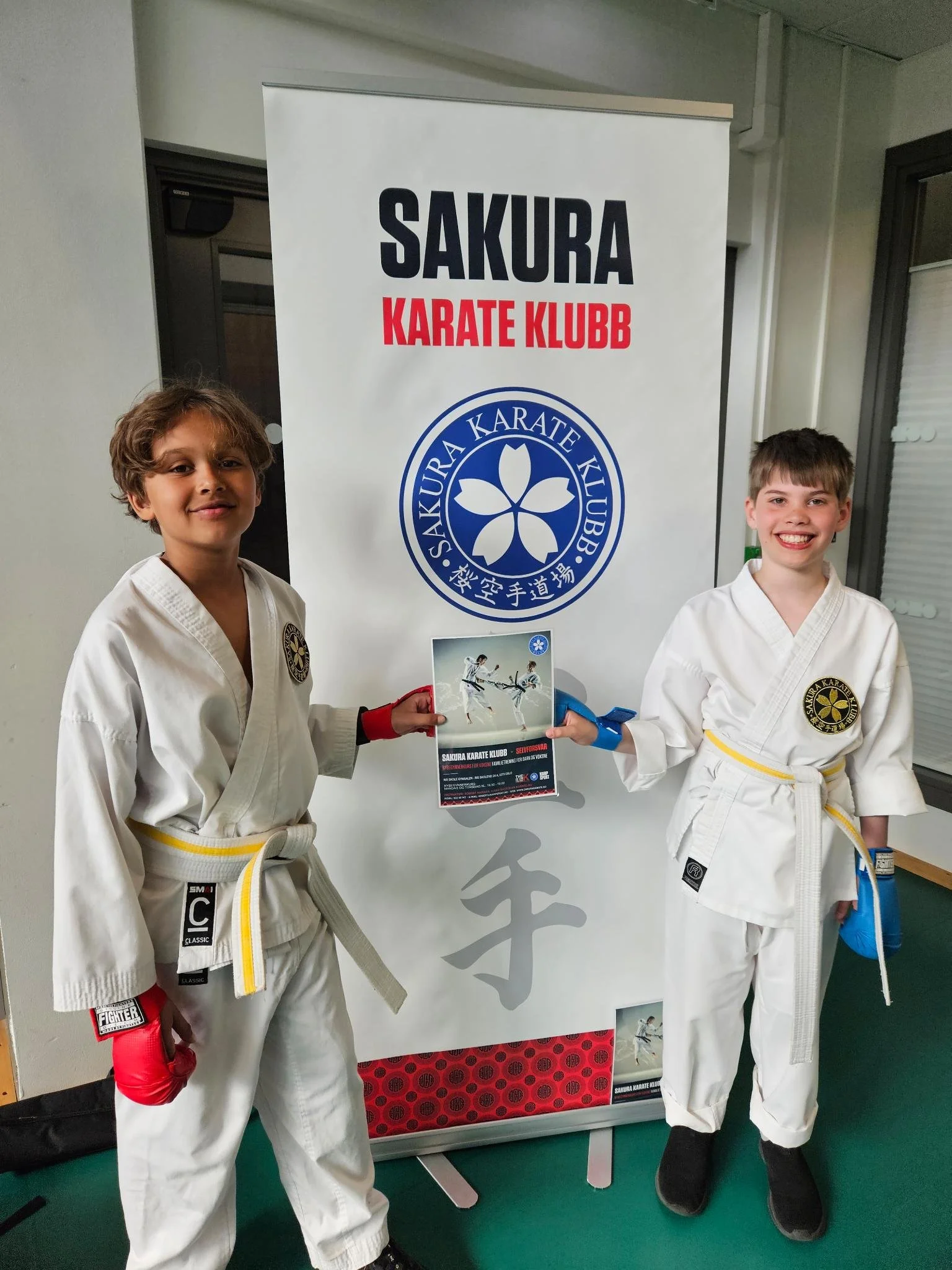 Stolte unge karateutøvere på aktivitetsdagen på Slemdal skole i hvite drakter står foran en banner for Sakura Karate Klubb, og holder en flyer. Begge smiler.