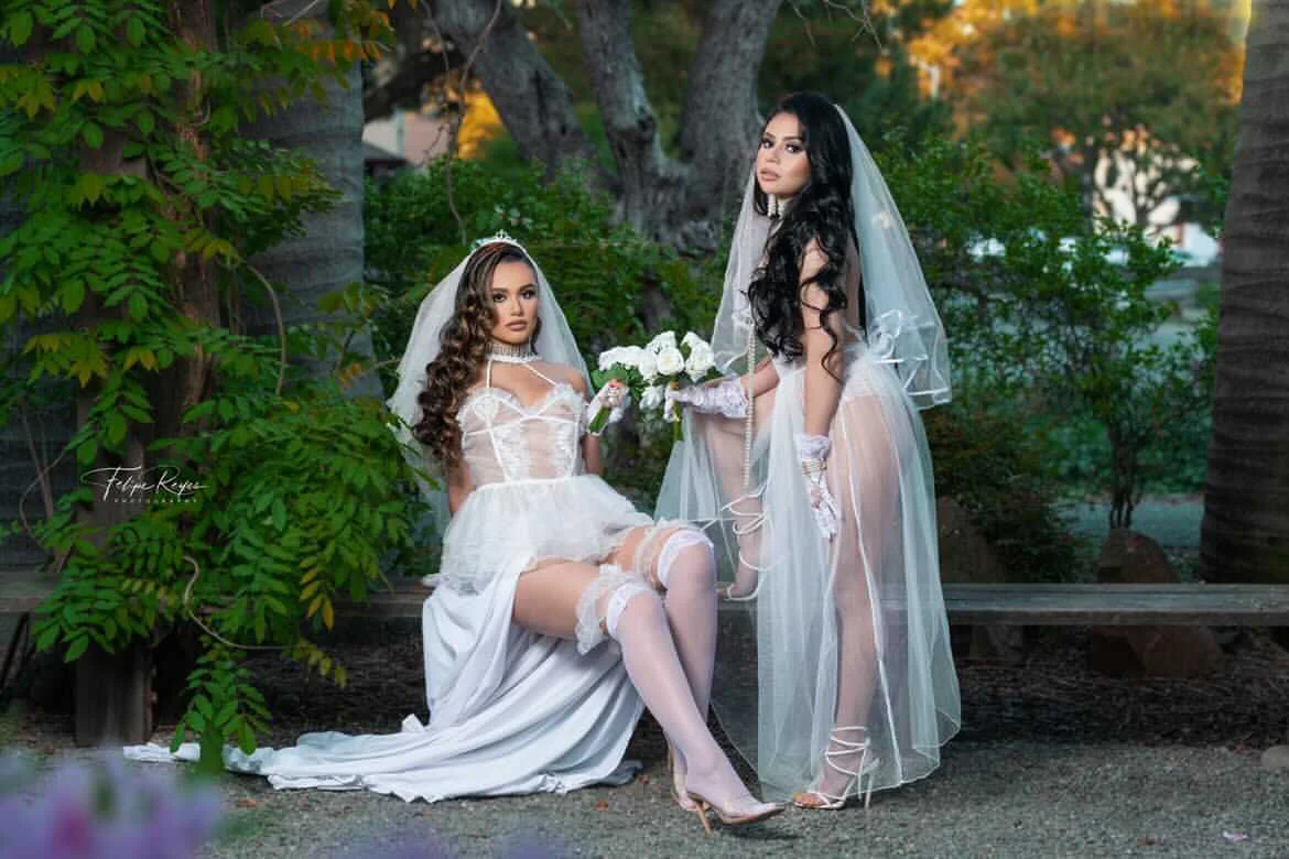 Two women dressed in bridal gowns with veils, one sitting and one standing, outdoors in a lush, green garden setting. One woman is holding a bouquet of white flowers.