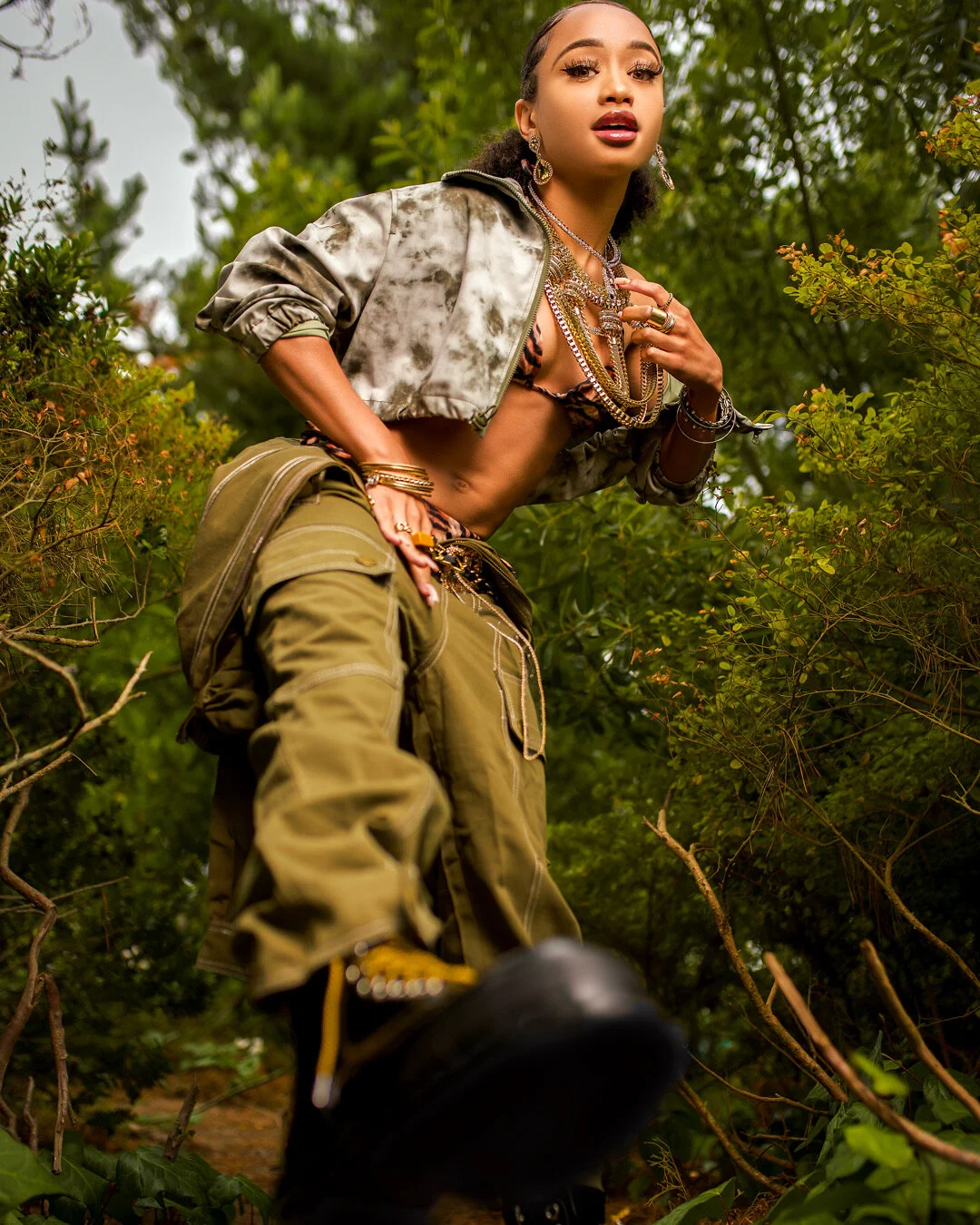 Young woman with jewelry, a partially open jacket, and cargo pants outdoors surrounded by green bushes and trees.