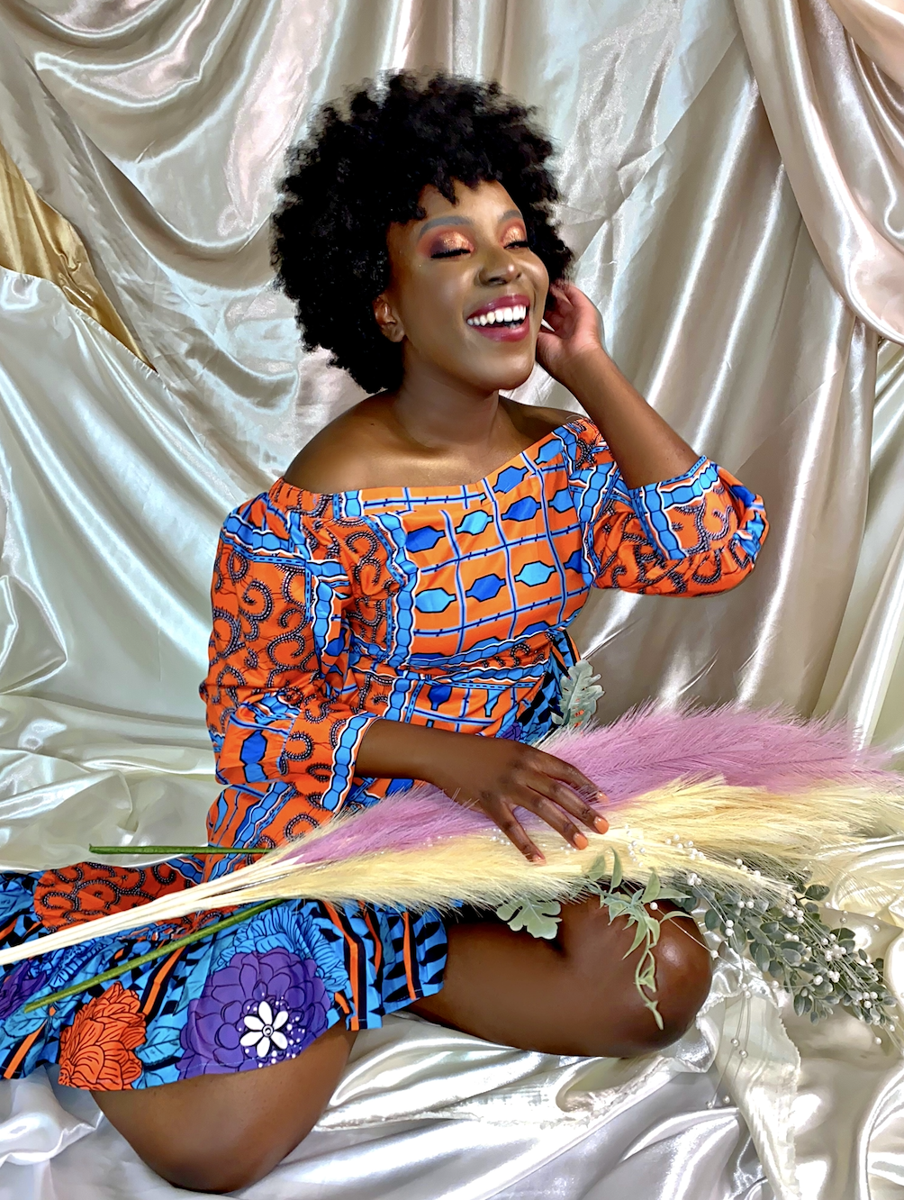 A woman with dark curly hair, smiling and dressed in a colorful, patterned off-shoulder dress, sitting on shiny fabric with a pink, yellow, and green floral arrangement in her lap.