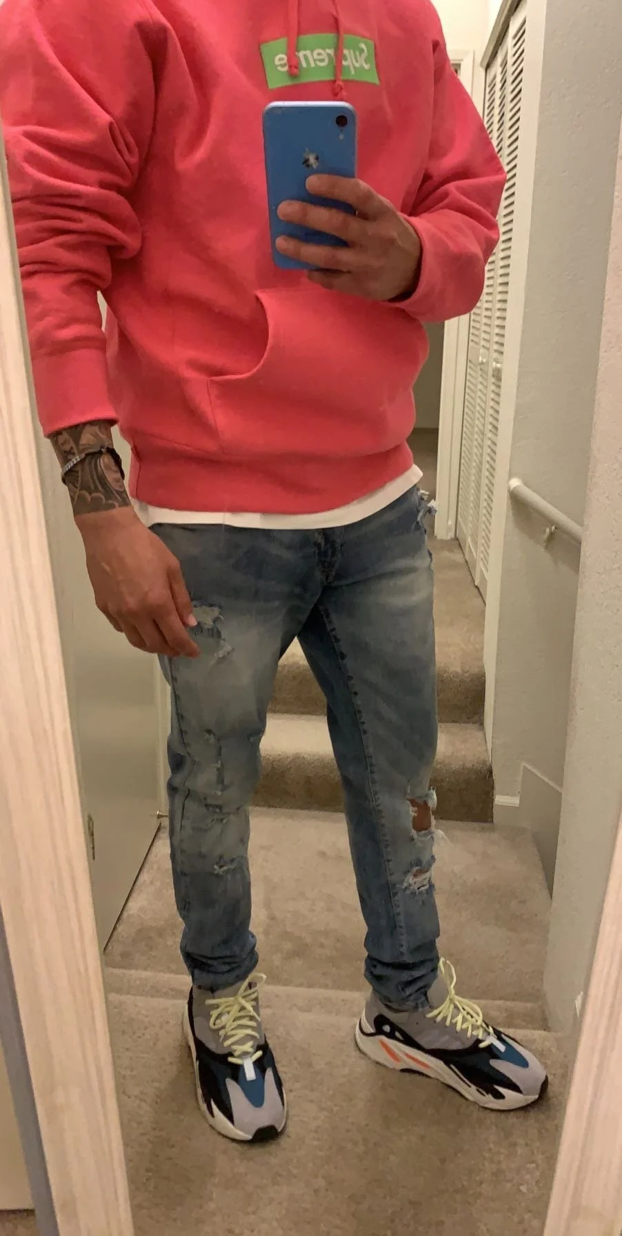 A person wearing a pink hoodie with a 'Supreme' logo, distressed blue jeans, and multi-colored sneakers, taking a mirror selfie in a room.
