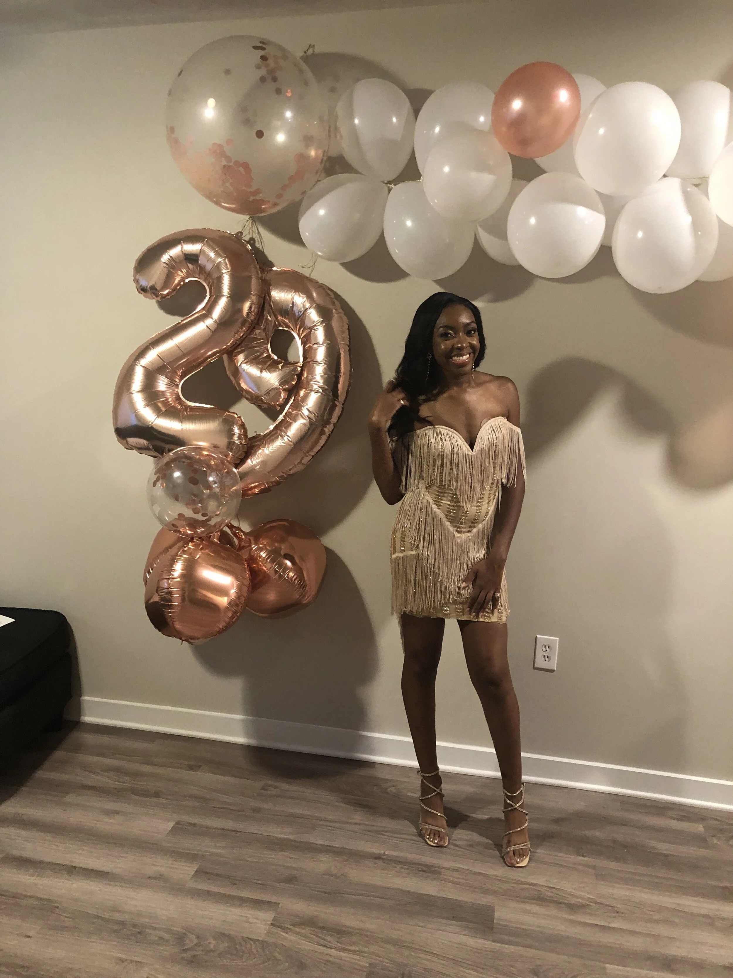 A woman in a gold dress standing indoors next to a decorative balloon arrangement, including a large rose gold number "22" balloon, white and rose gold balloons, and a transparent balloon with confetti.