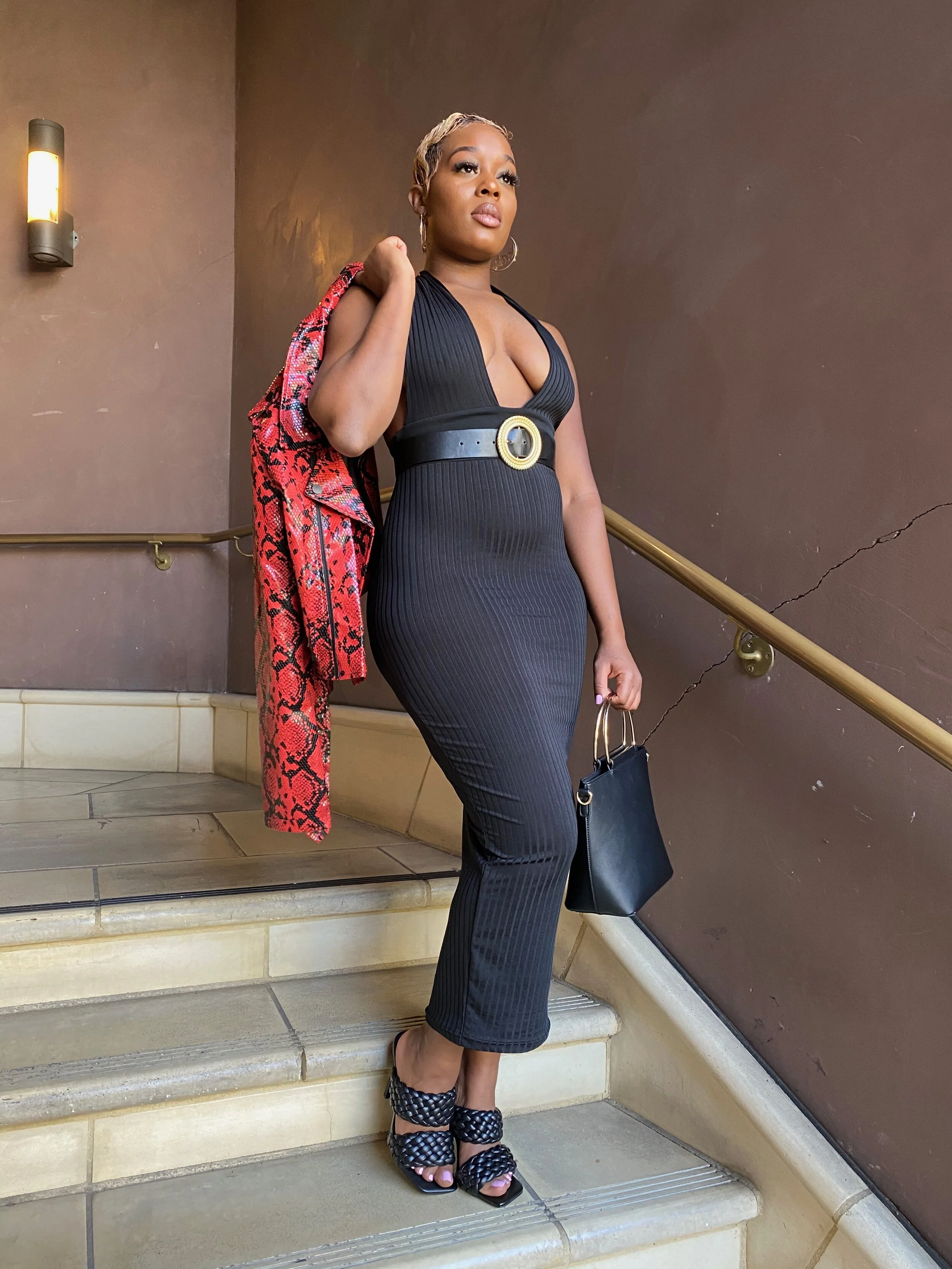 A woman dressed in a black sleeveless jumpsuit with a wide belt, holding a black handbag, and wearing black woven high-heeled sandals, standing on a staircase with a red patterned jacket over her shoulder, inside a building.