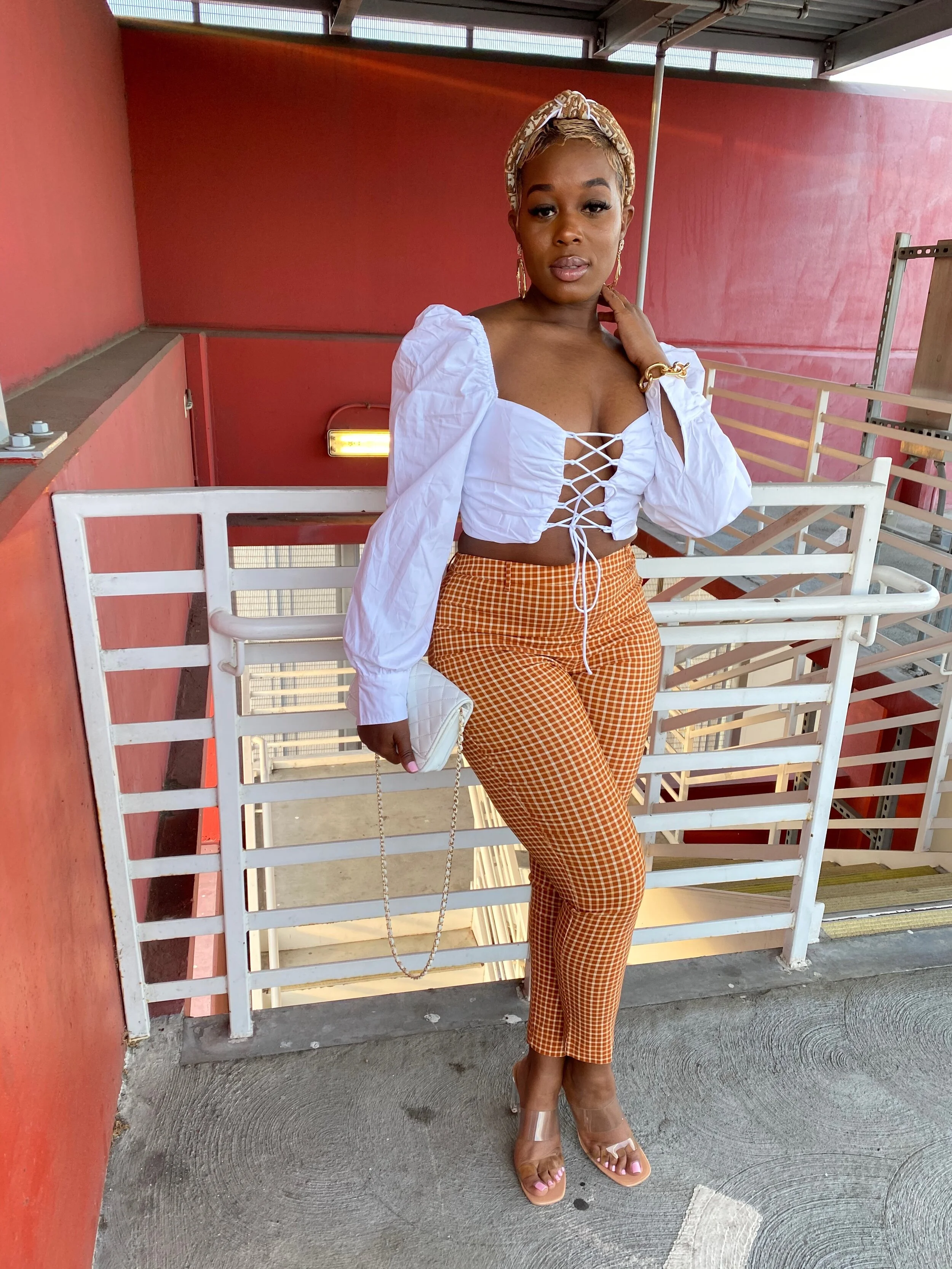 A woman standing outdoors on a balcony, wearing a white lace-up crop top, orange plaid pants, beige high heels, and accessories including earrings, a bracelet, a headband, and holding a small quilted purse.