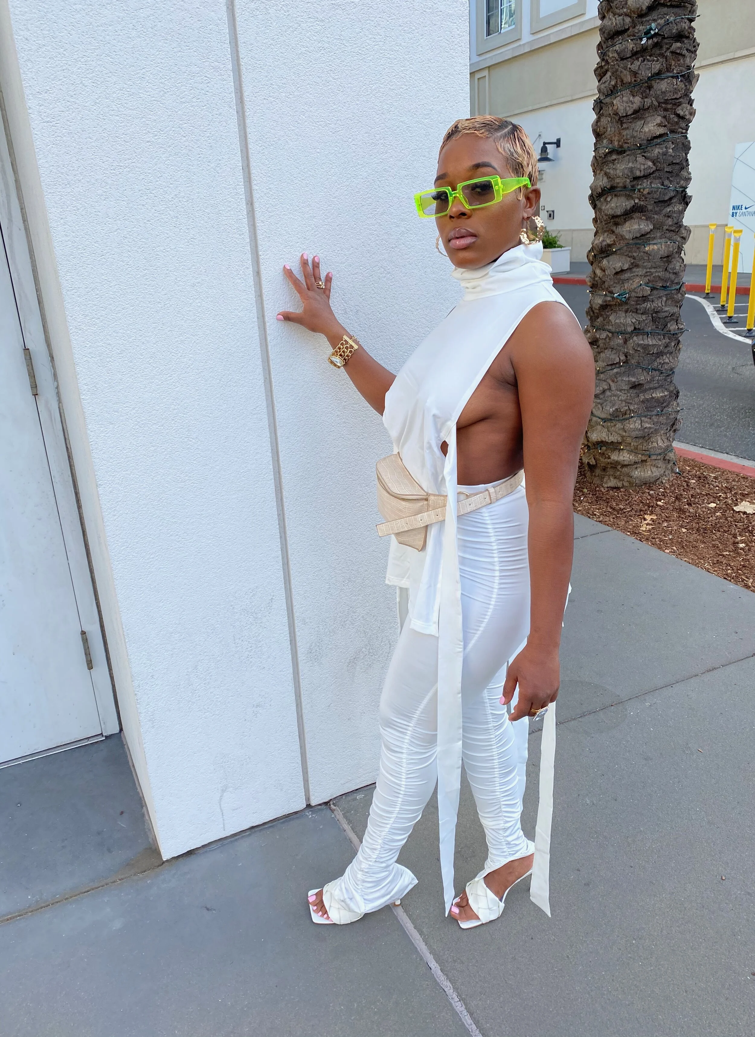 A woman in all white clothing and bright green sunglasses standing outdoors on a sidewalk, touching a white wall, with a palm tree and parked cars in the background.