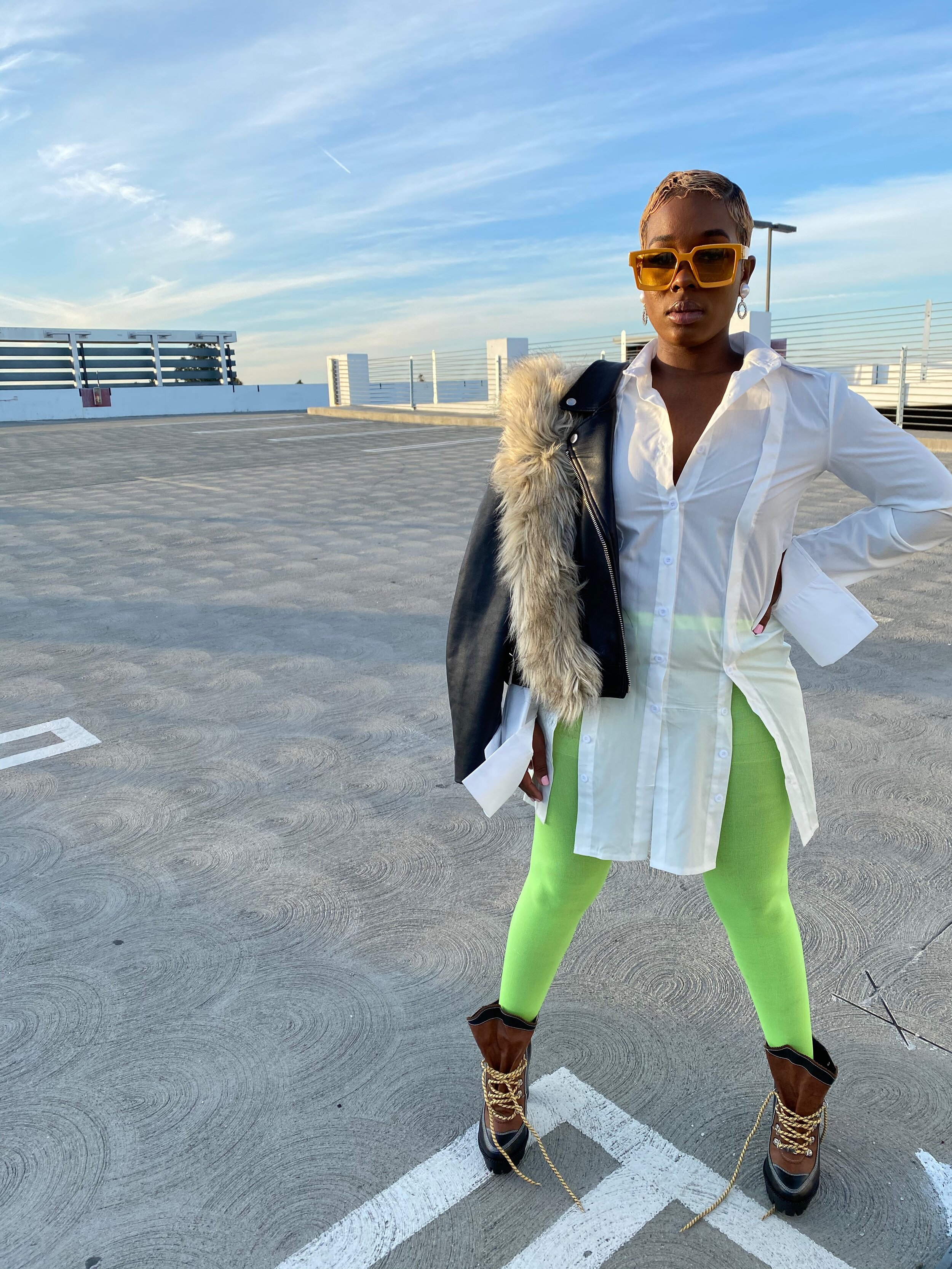 A woman standing in an empty outdoor parking lot, wearing oversized yellow sunglasses, a white button-up shirt, lime green leggings, and brown high-top boots with gold chains. She is holding a black jacket with a fur collar over her shoulder and has 