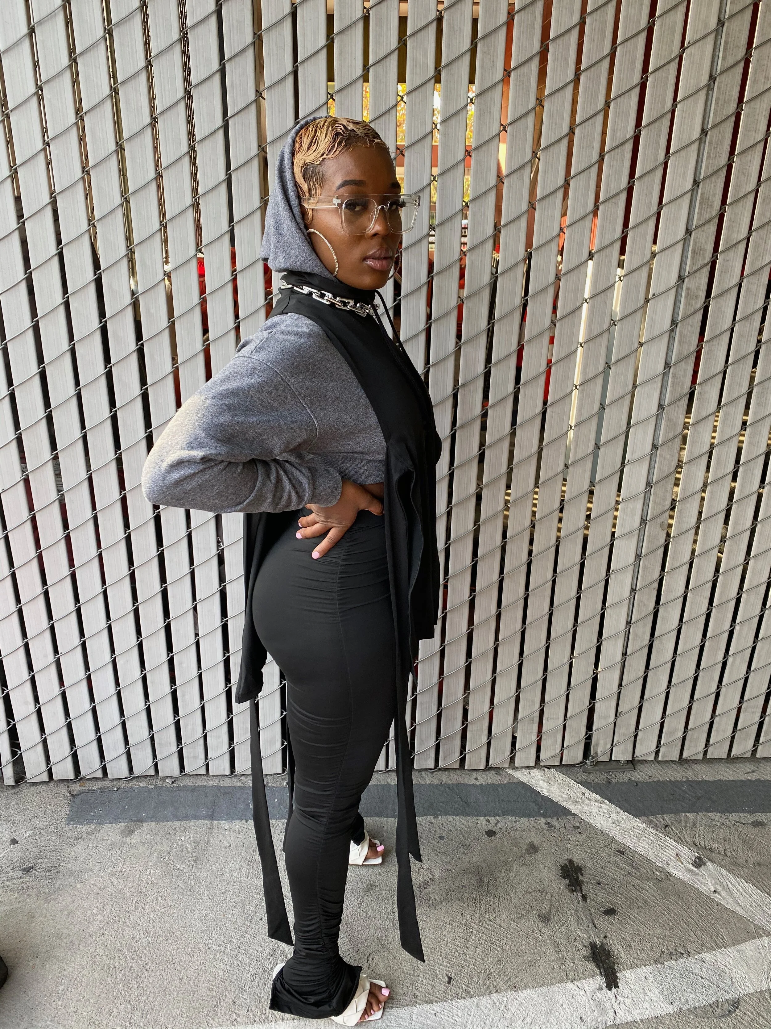 A woman standing outdoors in front of a wooden fence, wearing glasses, hoop earrings, a gray hoodie with a black vest, black leggings, and white high-heeled sandals.