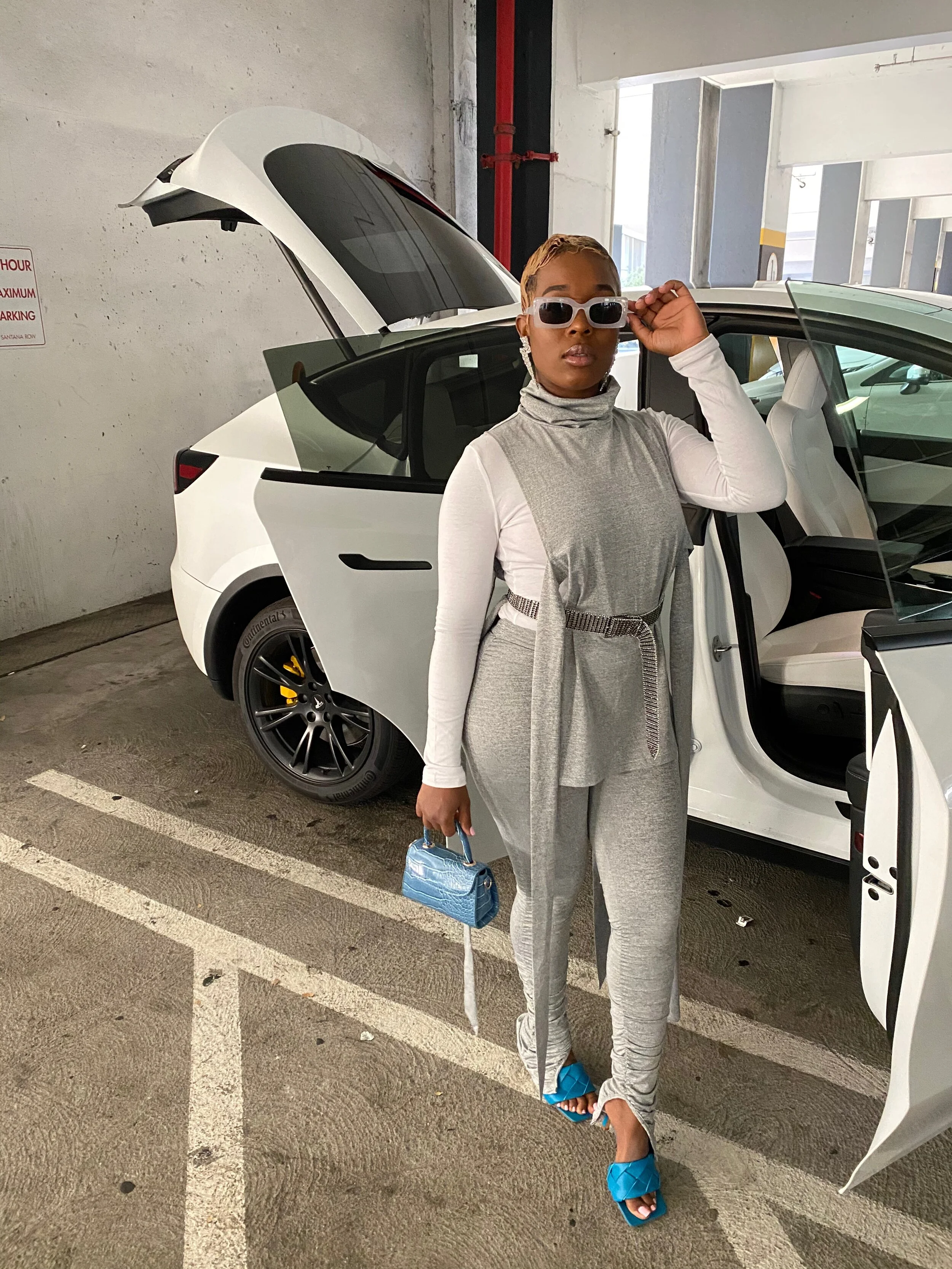Woman standing in a parking garage next to a white Tesla with the door open, wearing sunglasses, gray and white athletic wear, blue sandals, holding a small blue bag, and adjusting her sunglasses.