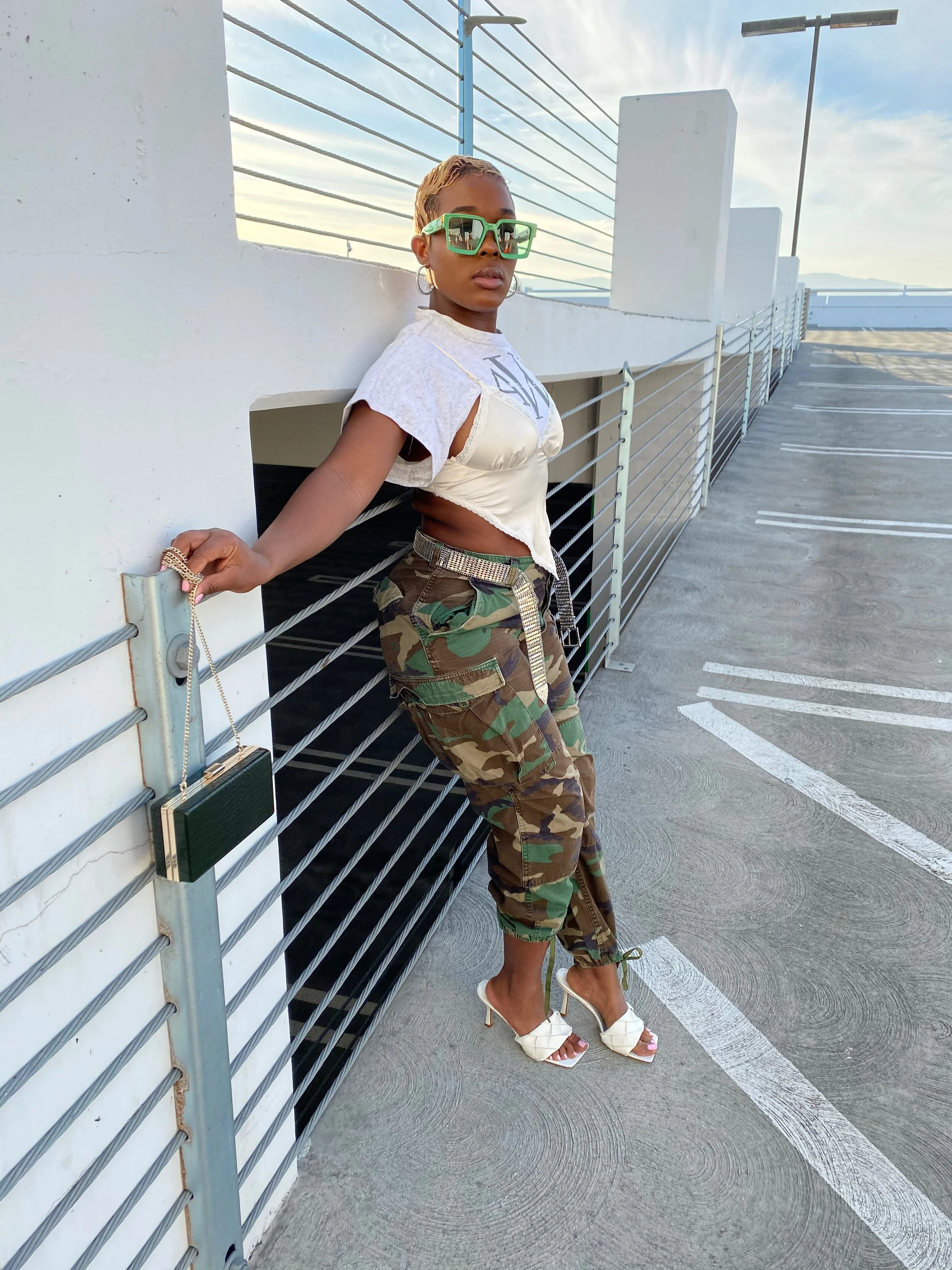 A woman leaning against a railing on a rooftop parking garage, wearing large green sunglasses, a white t-shirt with cutouts, camouflage cargo pants, and white high-heeled sandals.