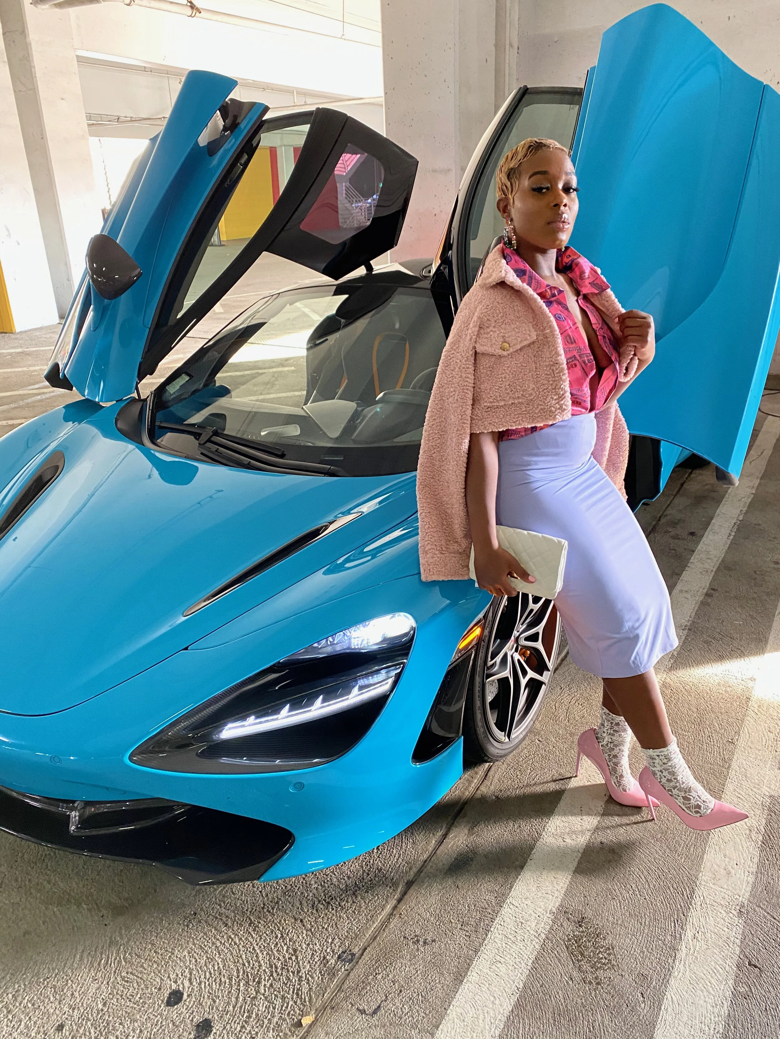 A woman standing beside a blue sports car with butterfly doors open, wearing a pink dress, pink high heels, lace socks, a light purple skirt, and a beige coat, holding a beige clutch.