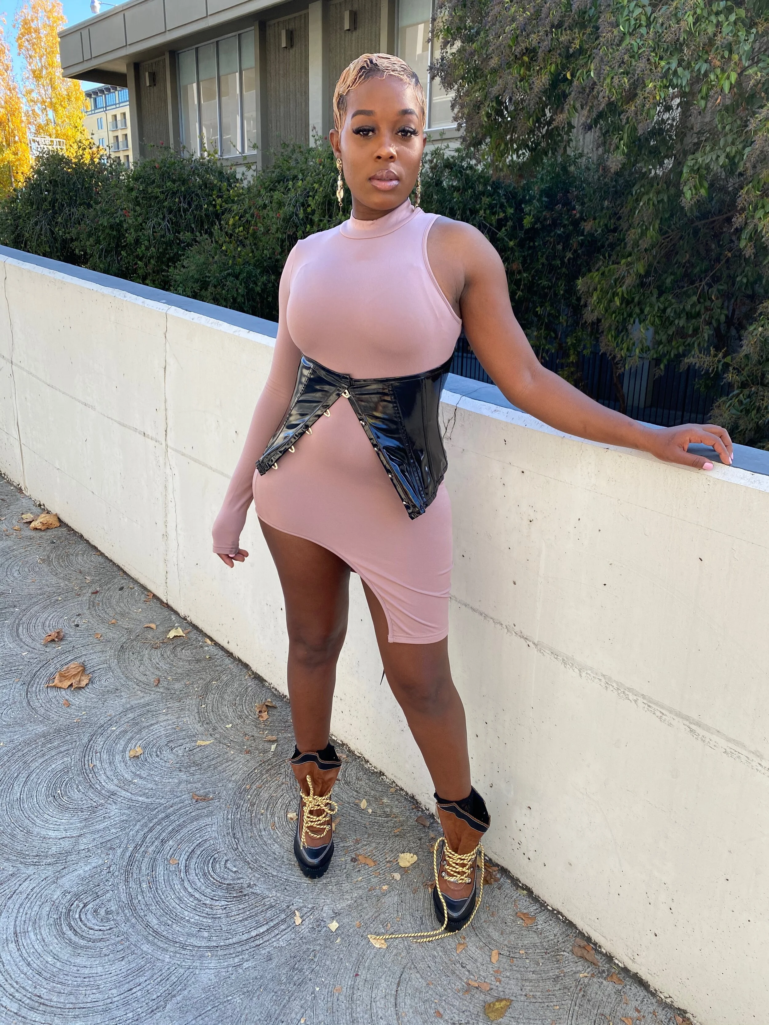 A woman in a pink dress, black patent leather waist trainer with gold eyelets, and black and gold high-heeled boots standing outside near a white concrete wall with greenery and a building in the background.