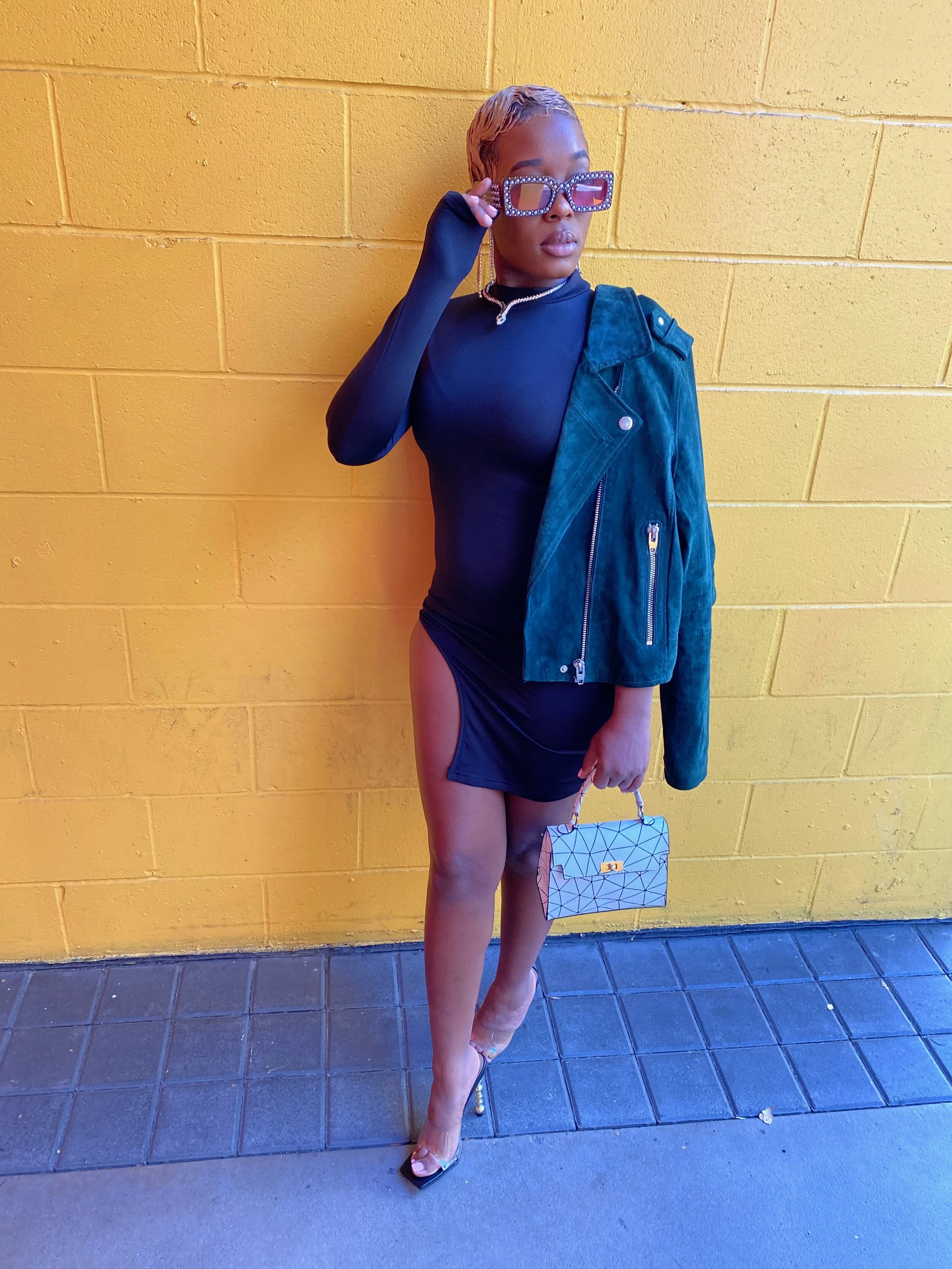 A woman standing against a yellow brick wall, wearing glasses, a black dress with side cutouts, a green jacket, high heels, and holding a patterned handbag.