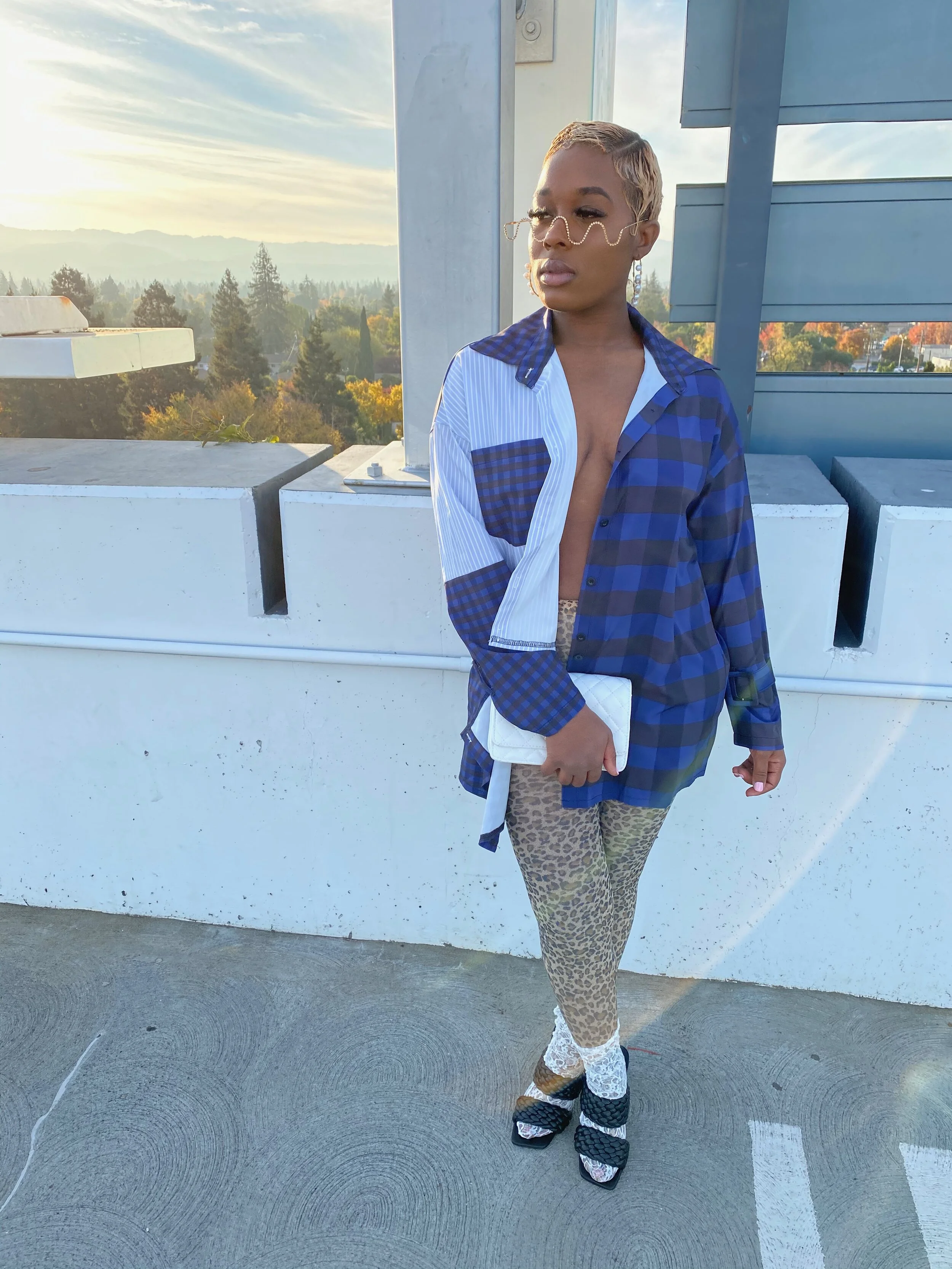 A woman standing on a rooftop parking lot, wearing a blue patchwork shirt, leopard print pants, and black high heels, with a cityscape and sunset in the background.