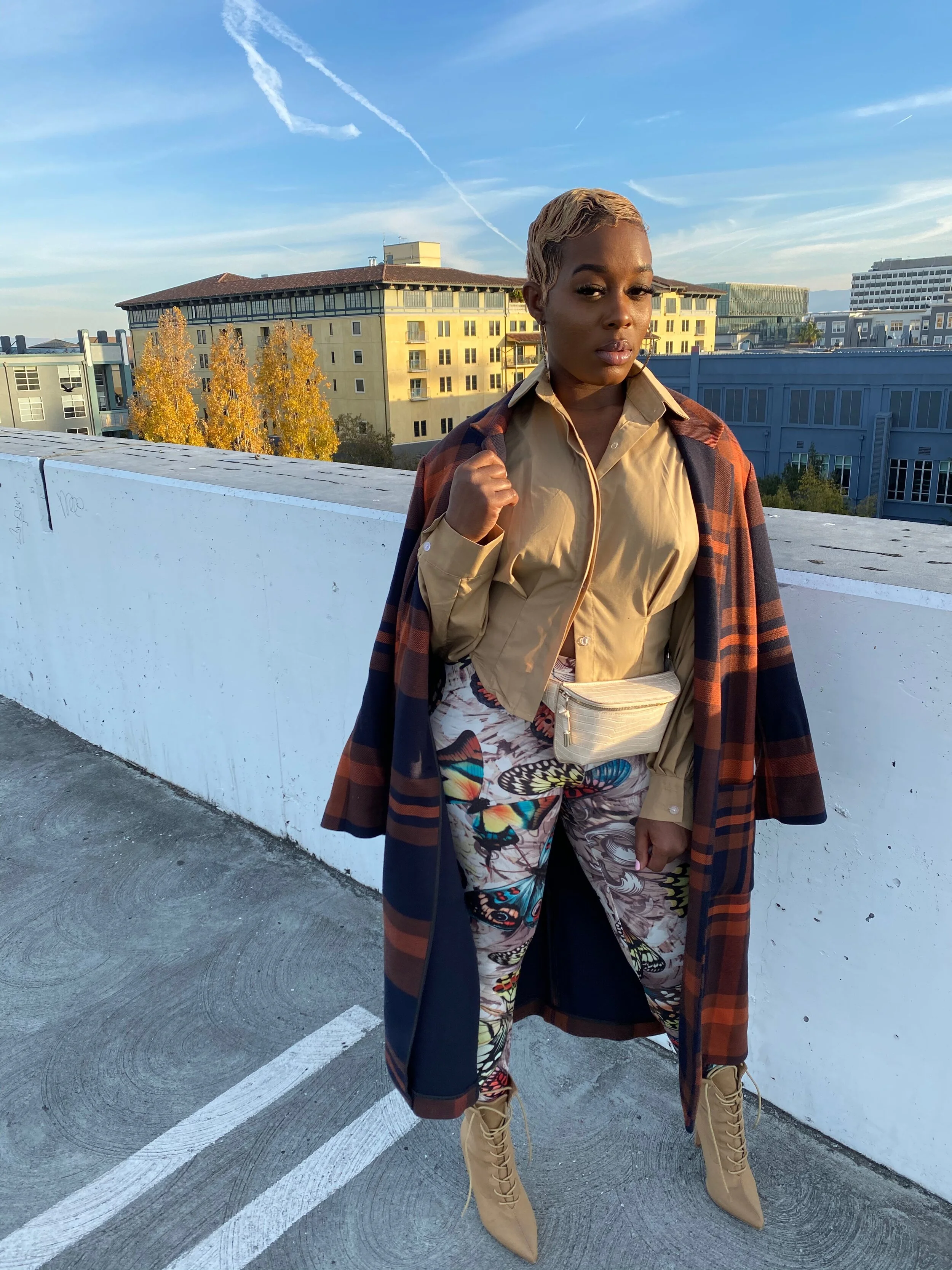 A woman standing on a parking garage rooftop, wearing a beige shirt, patterned pants, a long plaid coat, and beige lace-up boots, with city buildings in the background during sunset.