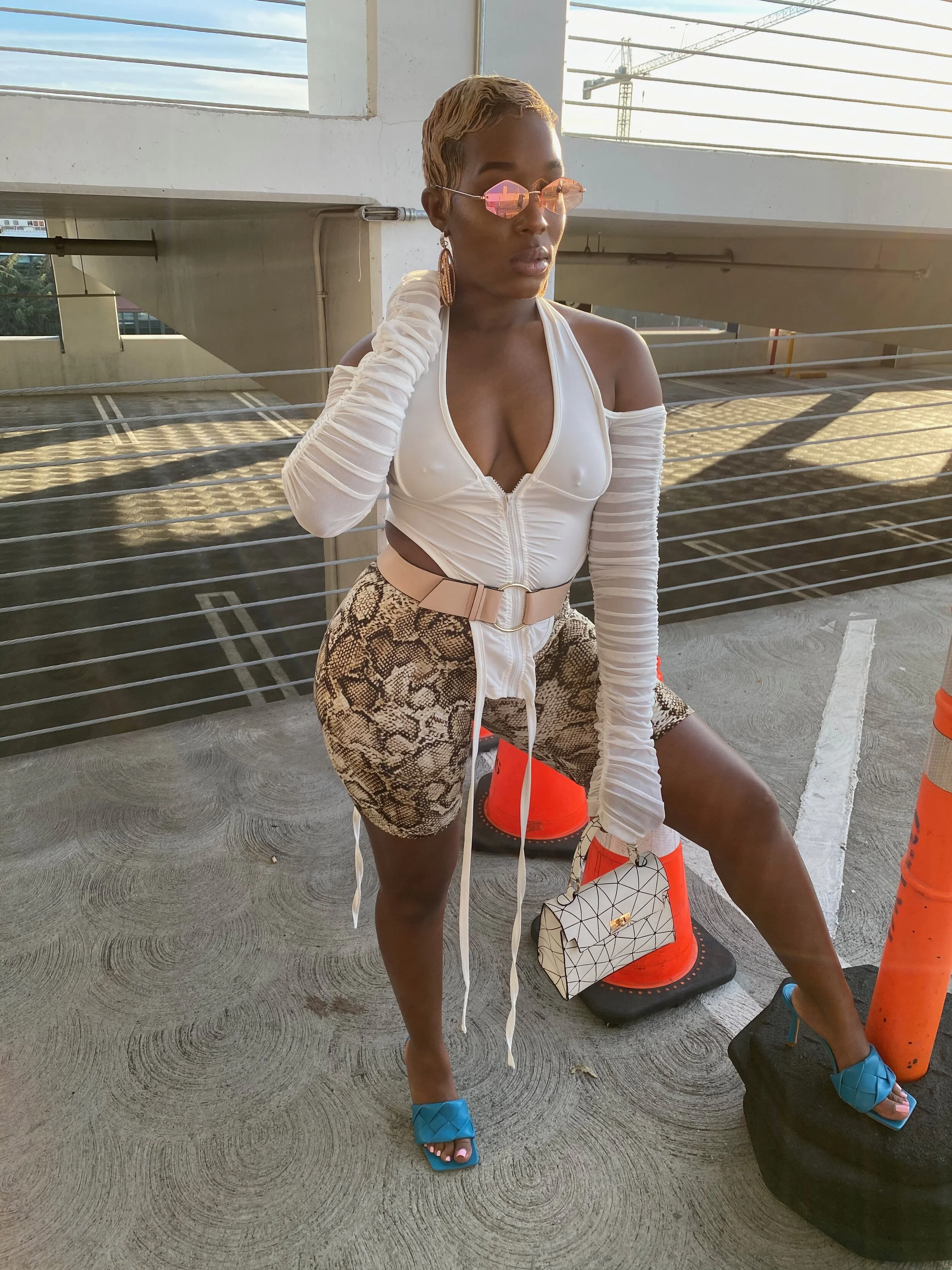 A woman with short blonde hair wearing pink oval sunglasses, a white zip-up top with off-shoulder sleeves, snake print shorts, blue high-heeled sandals, and holding a small geometric-patterned handbag. She is standing outdoors near a parking garage w