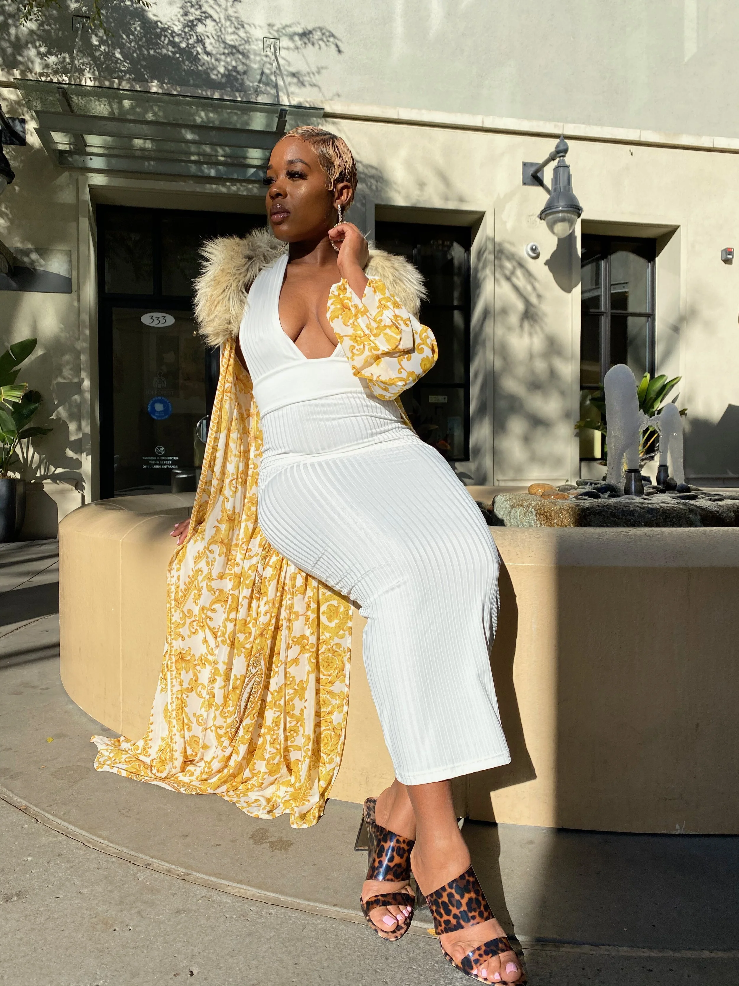 A woman wearing a white ribbed dress, leopard print high heels, and a patterned yellow and white coat with fur collar, sitting on a low beige wall outdoors in sunlight.
