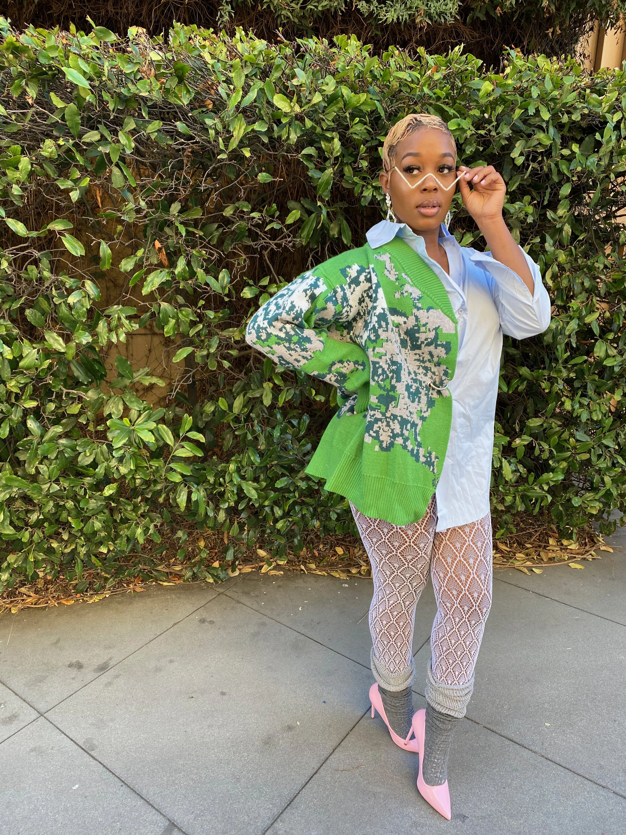 A woman standing outdoors in front of green bushes, wearing a white shirt dress, a green and gray patterned cardigan, fishnet tights, gray leg warmers, and pink high-heeled shoes, with long earrings and a chain across her face.