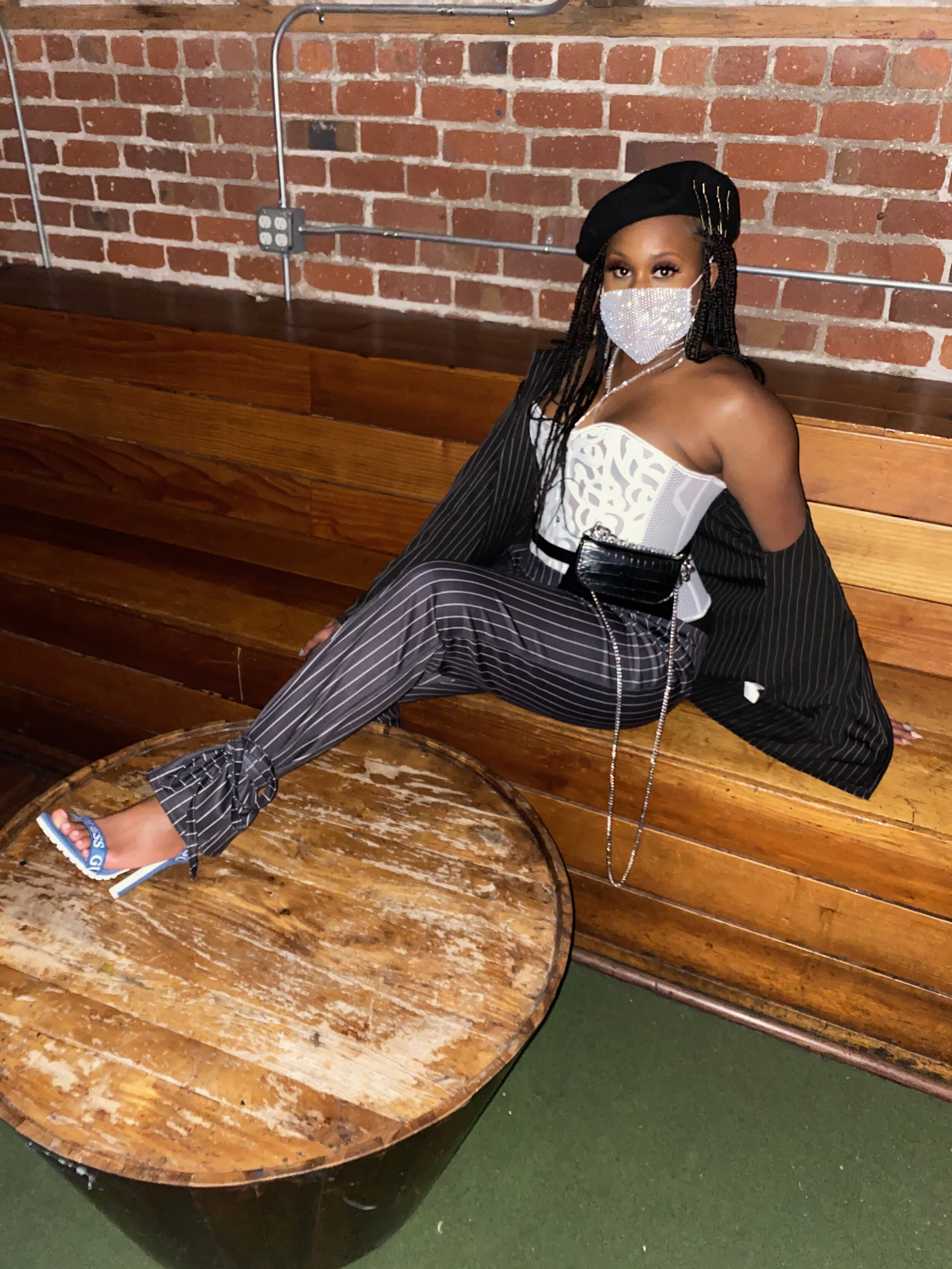 A woman sitting on a wooden bench in an indoor setting with a brick wall background, wearing a stylish off-shoulder top with a lace pattern, pinstriped pants, high heels, black beret, and a rhinestone face mask.