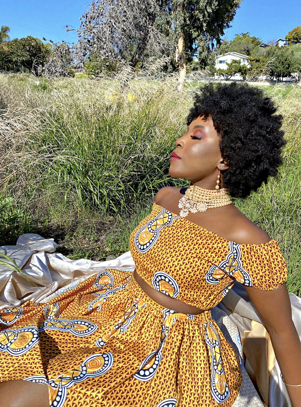 A woman with a natural curly hairstyle wearing colorful traditional African attire and jewelry, peacefully sitting outdoors with her eyes closed amidst tall grass and trees under a clear blue sky.