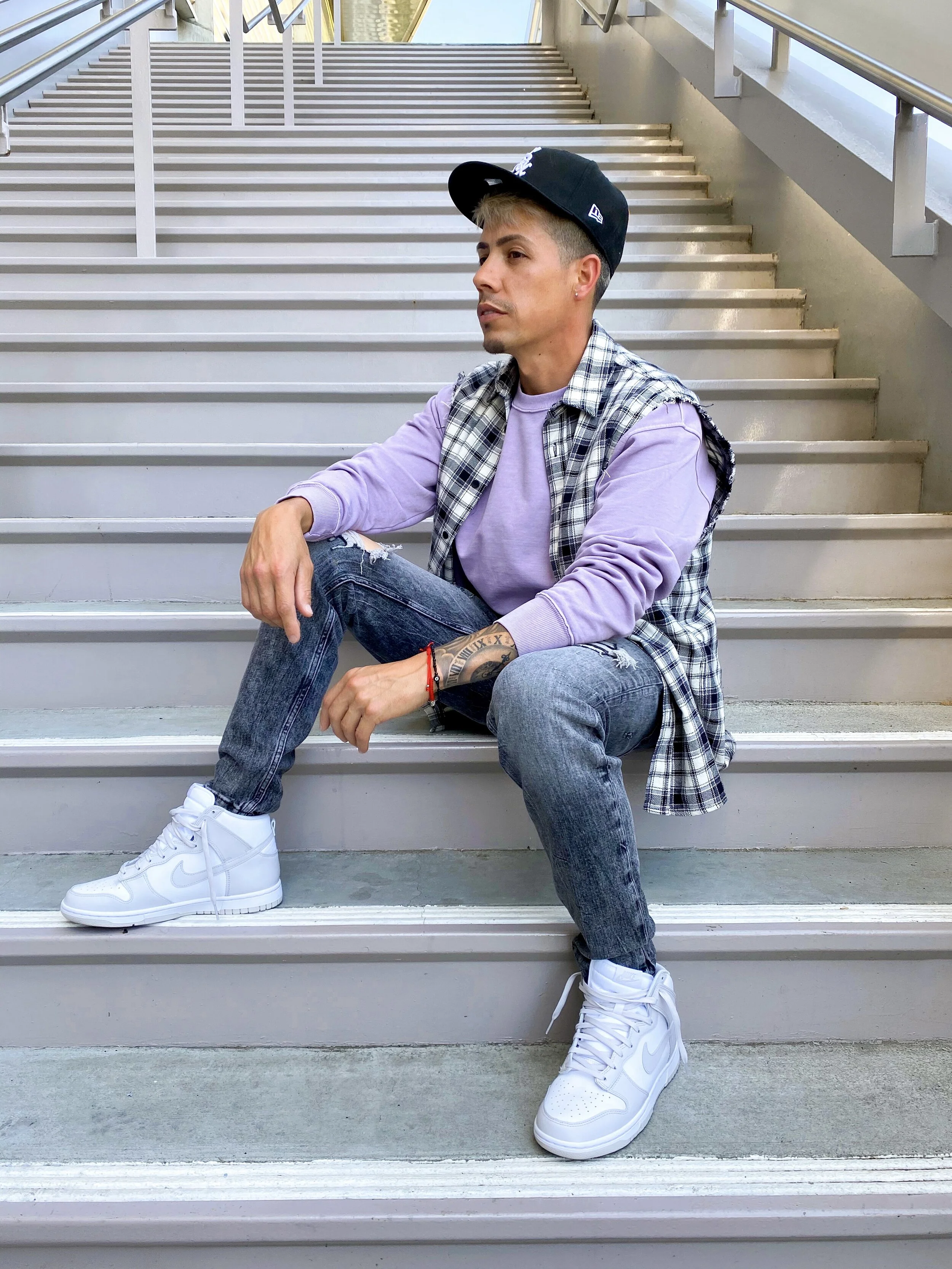 A young man with tattoos and short hair sitting on outdoor stairs wearing a black baseball cap, lavender long-sleeve shirt, plaid vest, ripped jeans, and white Nike sneakers.