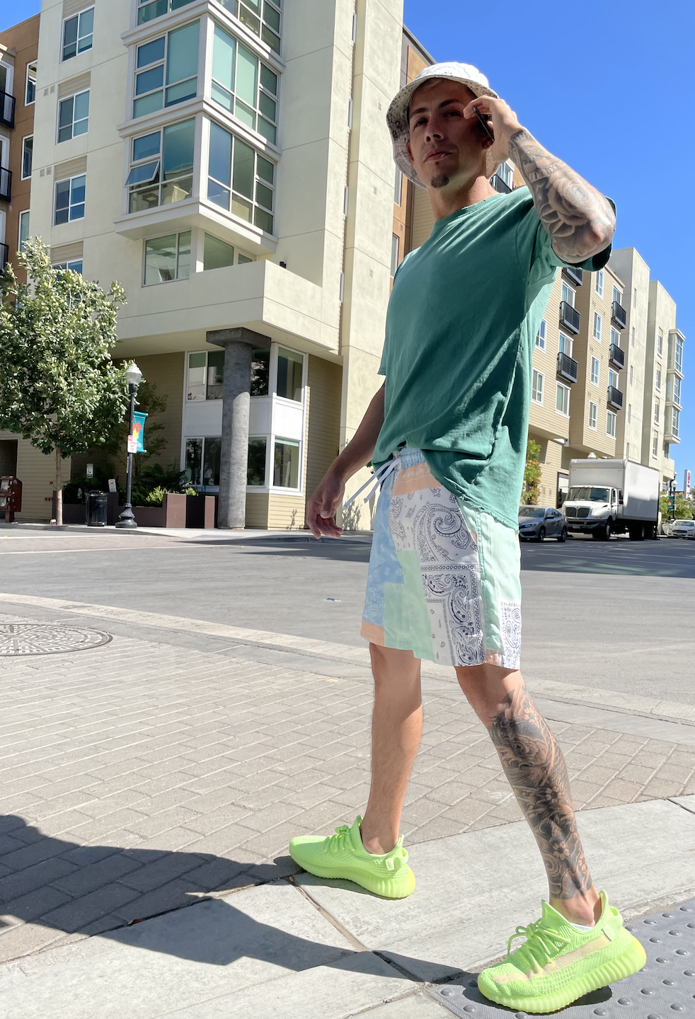Young man wearing a green t-shirt, colorful shorts, and neon green sneakers, talking on a cell phone, standing on a city sidewalk with modern apartment buildings and a clear blue sky behind him.
