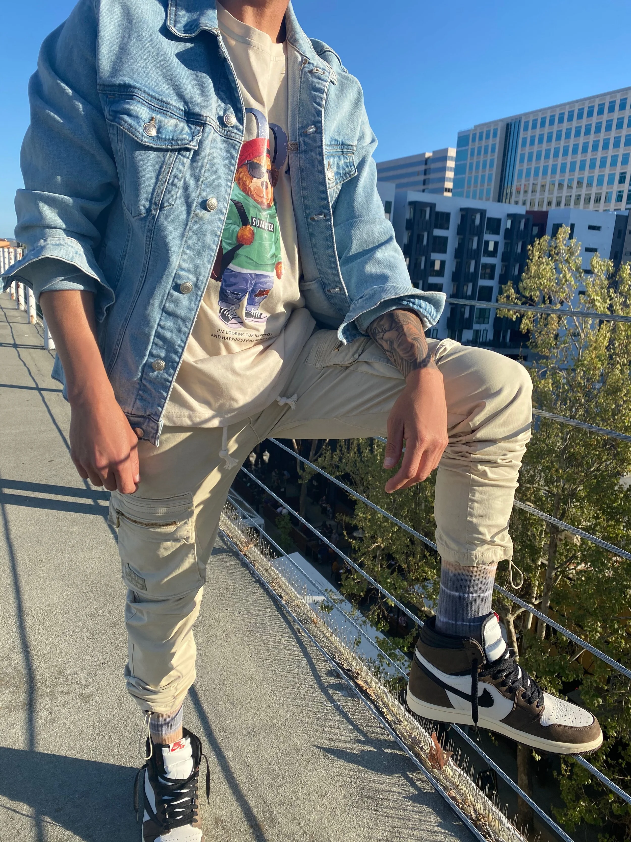 Person wearing a denim jacket, graphic t-shirt, beige jogger pants, and Nike Air Jordan sneakers, sitting on a rooftop ledge with an urban cityscape in the background.