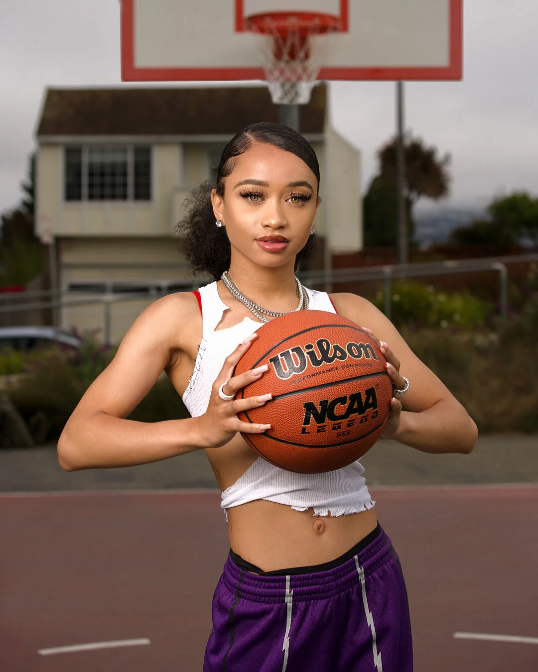 A young woman with earrings, a necklace, and rings, wearing a tattered white tank top and purple shorts, holding a basketball on an outdoor court with a backboard and hoop, and a house in the background.
