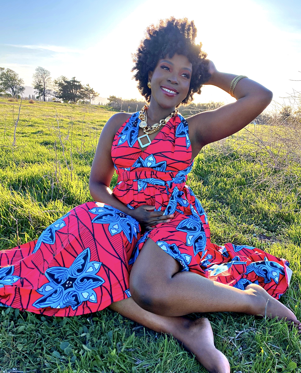 A woman sitting on green grass in a field during sunset, wearing a colorful red dress with blue and black patterns, smiling with her hand resting on her head, accessorized with gold jewelry.