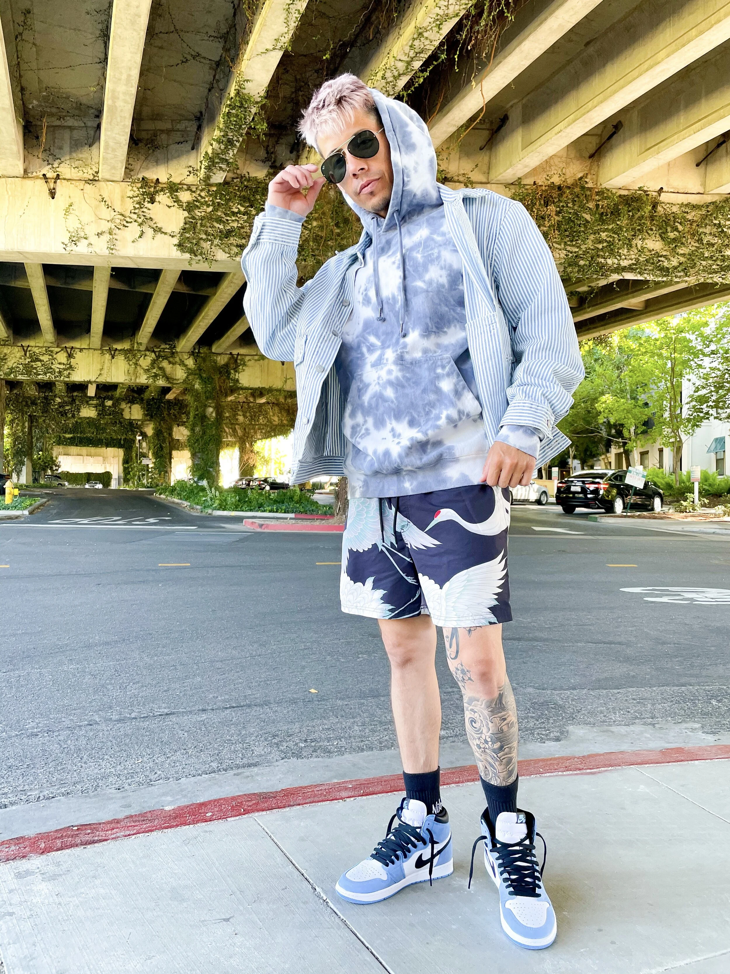 A young man with blond hair wearing sunglasses, a tie-dye hoodie, a striped jacket, swim shorts with cranes, and sneakers, standing under an overpass on a city street.