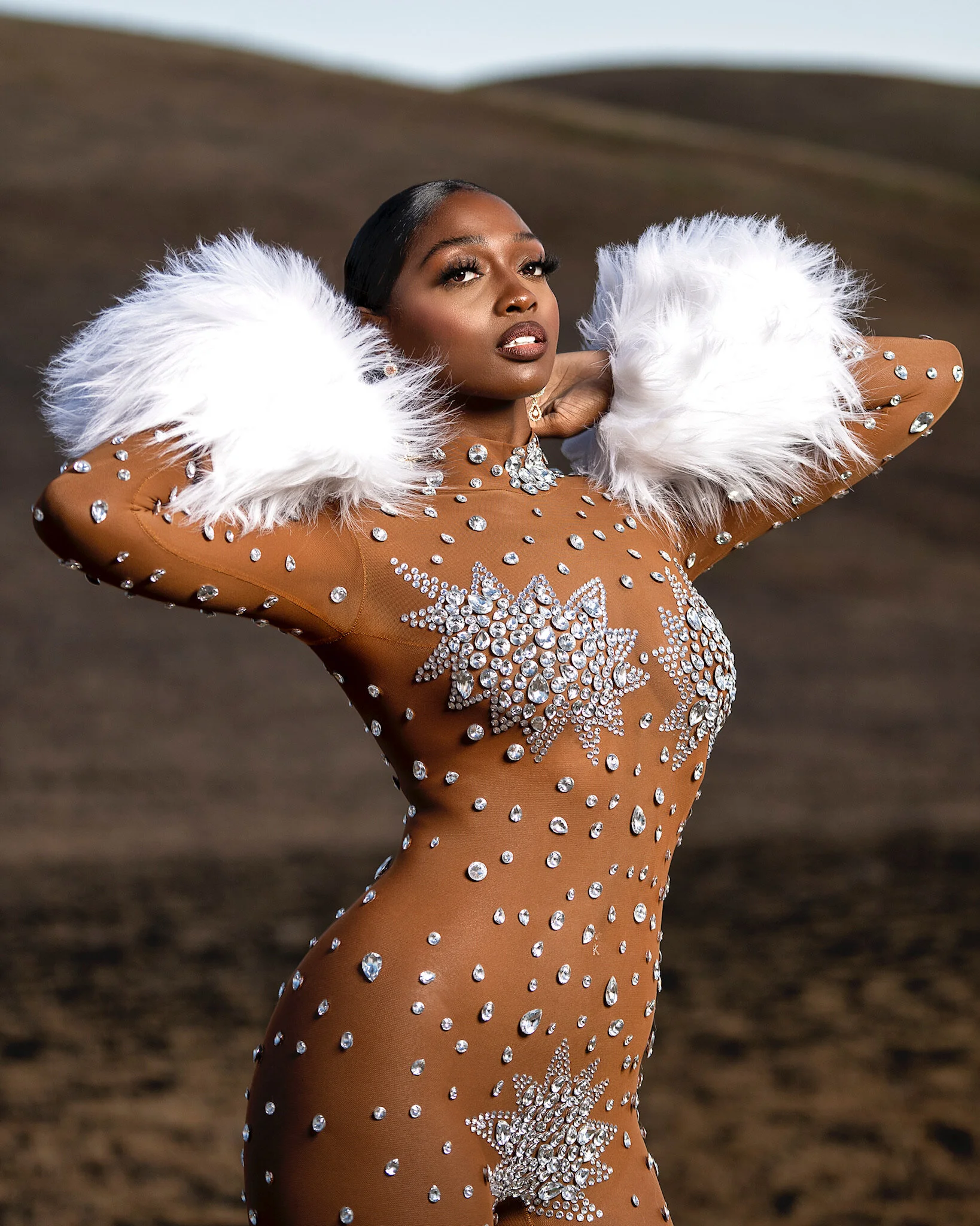 A woman wearing a form-fitting bronze-colored dress decorated with numerous rhinestones, with large white feathered sleeves and a choker necklace, standing outdoors in a landscape with hills.