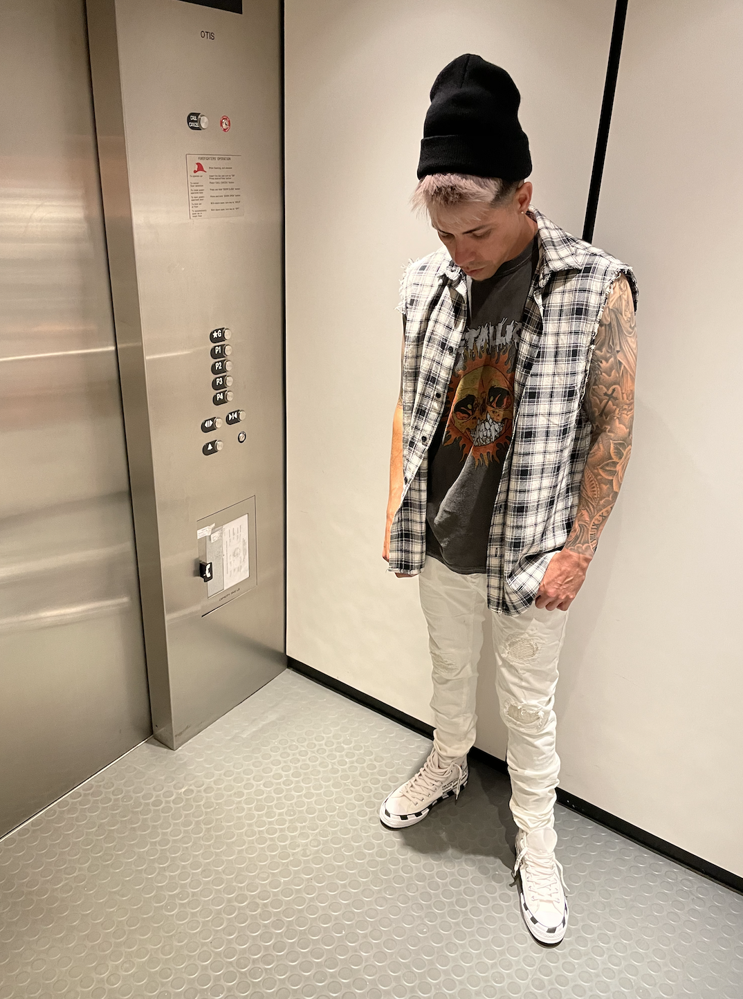 A person with pink hair wearing a black beanie, a sleeveless plaid shirt, a graphic T-shirt, distressed white jeans, and high-top sneakers, standing inside an elevator with one hand in their pocket, looking down.