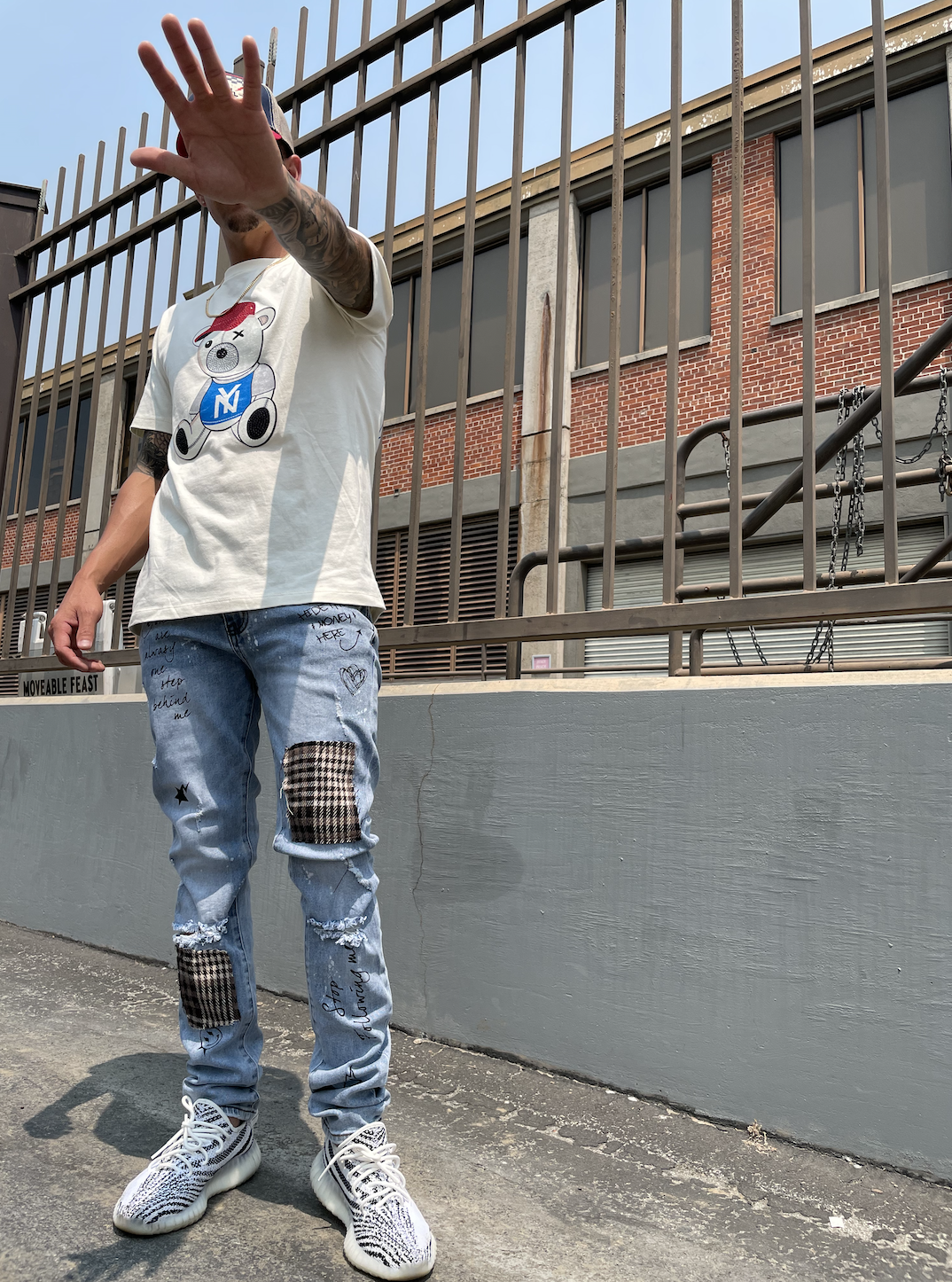 A person standing outdoors on a paved area near a gray wall, with a brick building and metal fence in the background. The individual is wearing a white t-shirt with a cartoon bear graphic, light blue ripped jeans with patches and handwritten notes, a