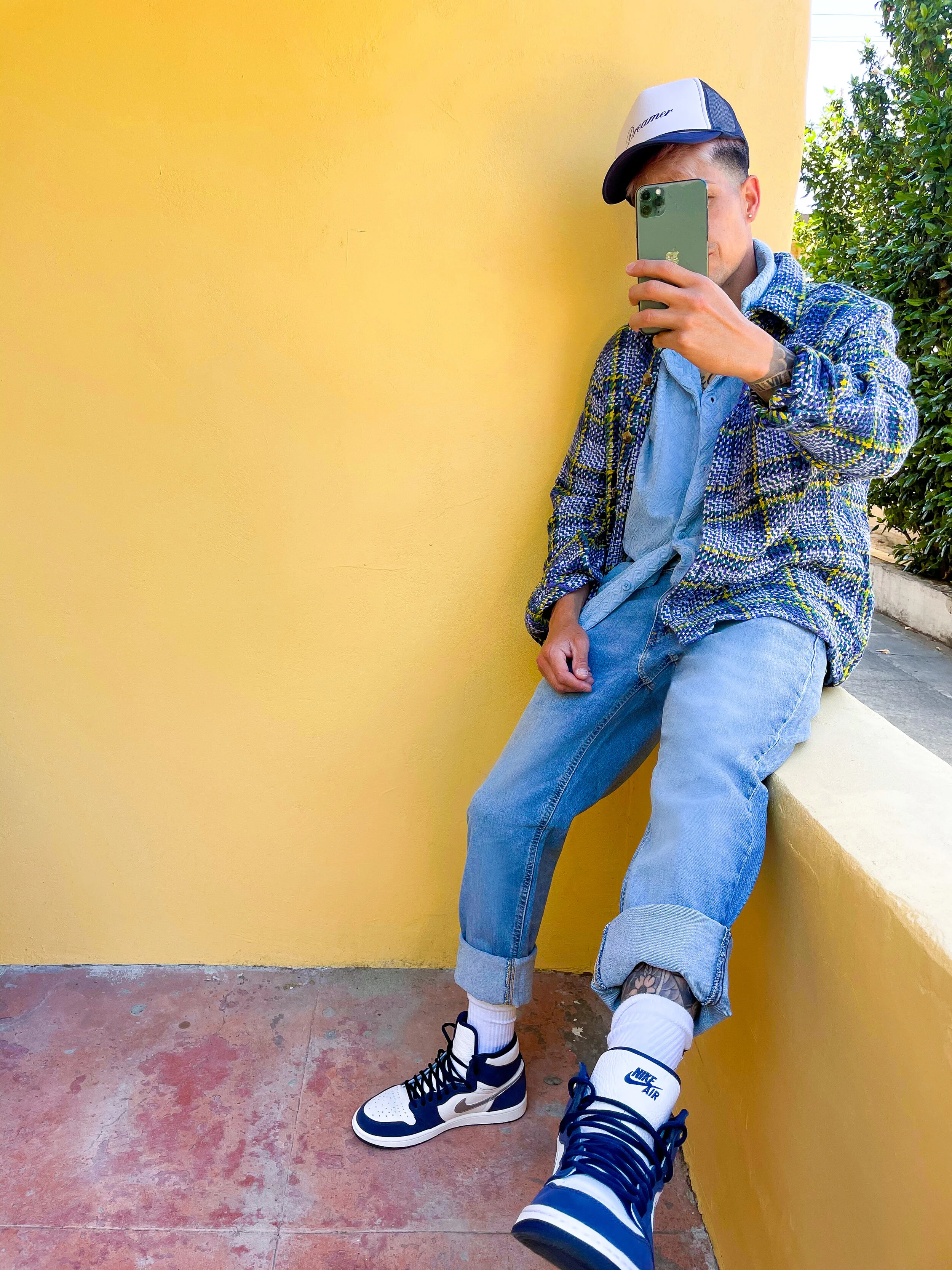 A person sitting on a yellow ledge taking a mirror selfie with their phone, wearing a cap, plaid jacket, jeans, and Nike sneakers.