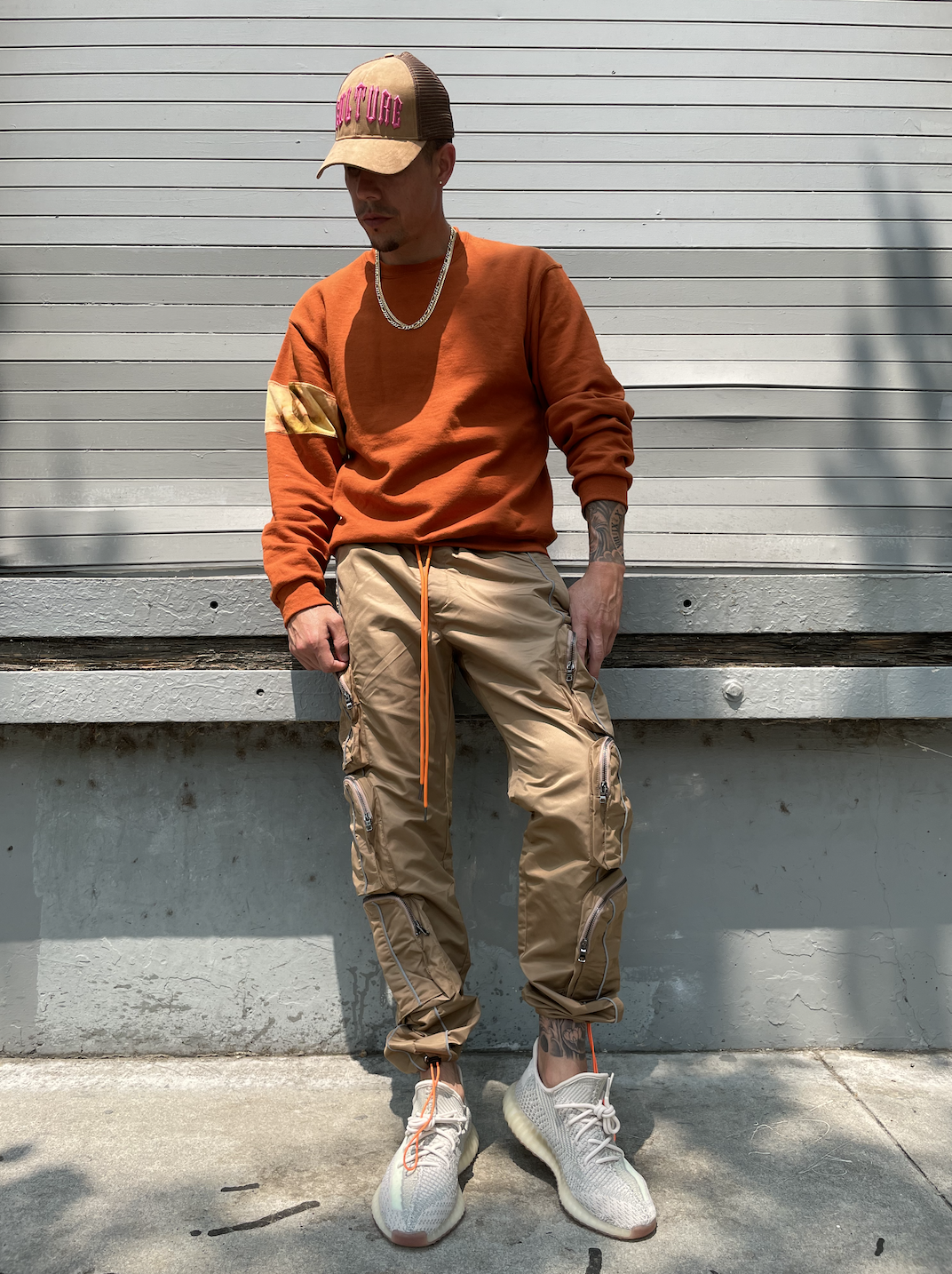 A man wearing a beige cap with pink text, an orange sweatshirt, beige cargo pants, and white sneakers standing against a white garage door.