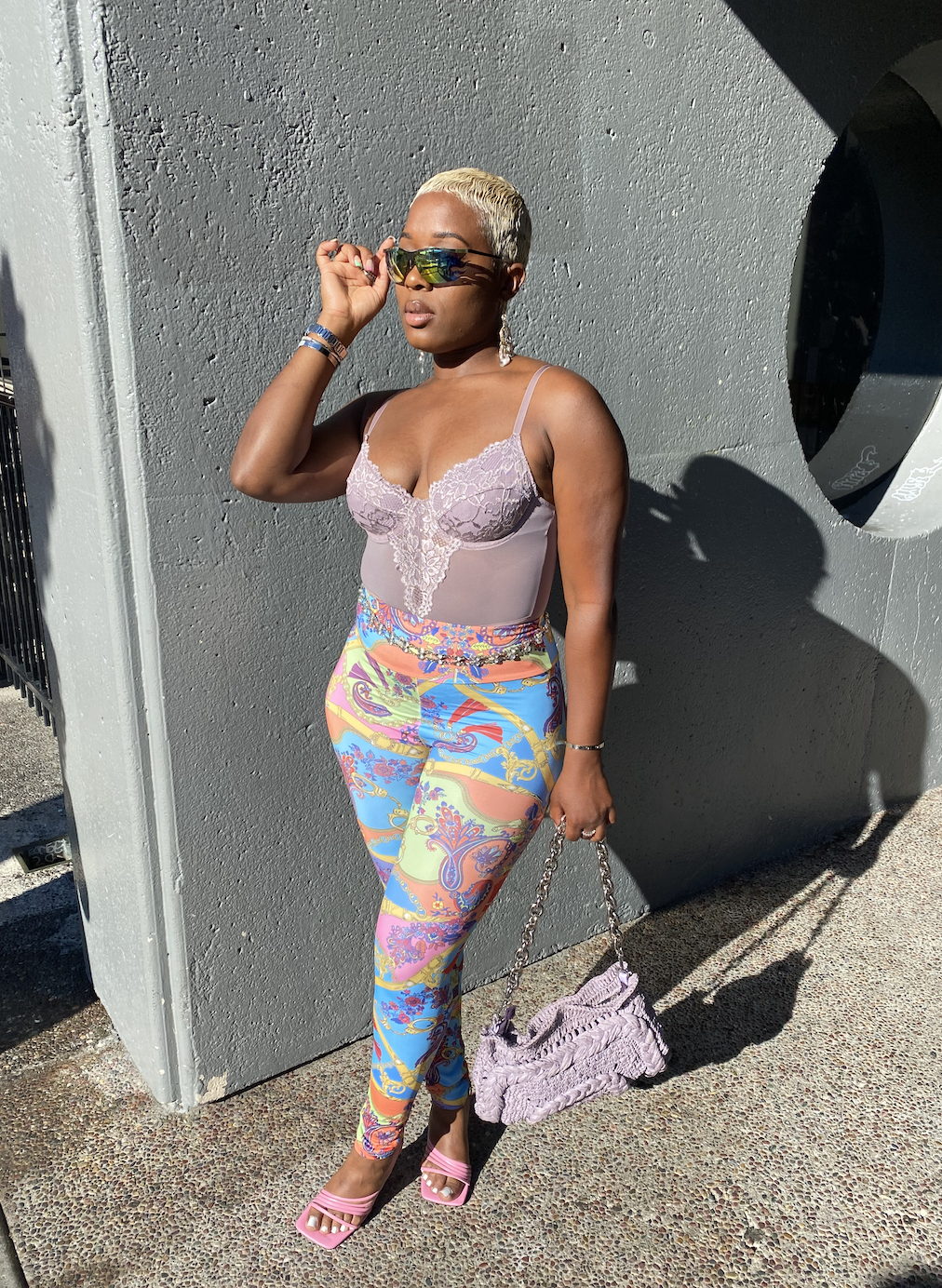 A woman standing against a gray wall, wearing colorful patterned pants, a light pink lace top, pink high heels, and holding a purple handbag, with sunglasses pushed up on her head.