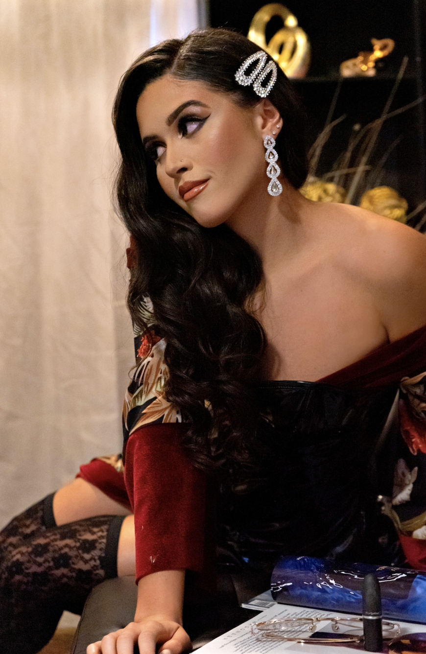 A woman with styled black hair and makeup, wearing large diamond earrings and a decorative hair clip, sits at a table with jewelry and makeup products, indoors with a dark background and decorative items.