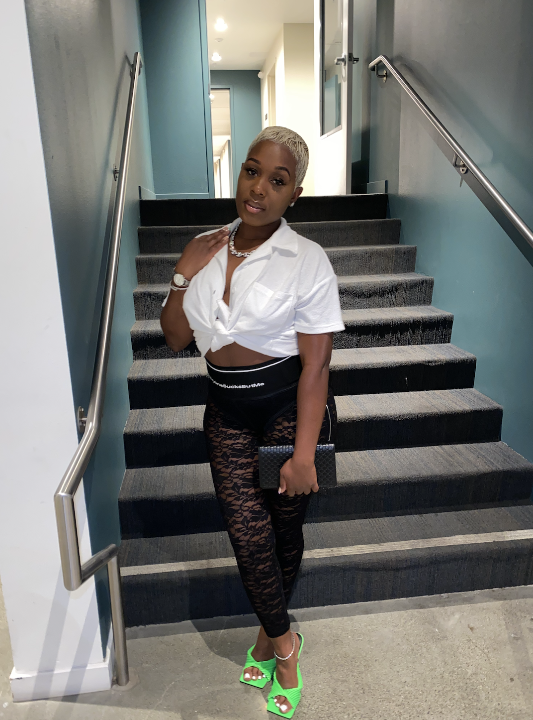 A woman with short platinum blonde hair standing on a staircase, wearing a white knotted crop top, black lace leggings, neon green high heels, and accessories including a watch, bracelet, necklace, and holding a small black purse.