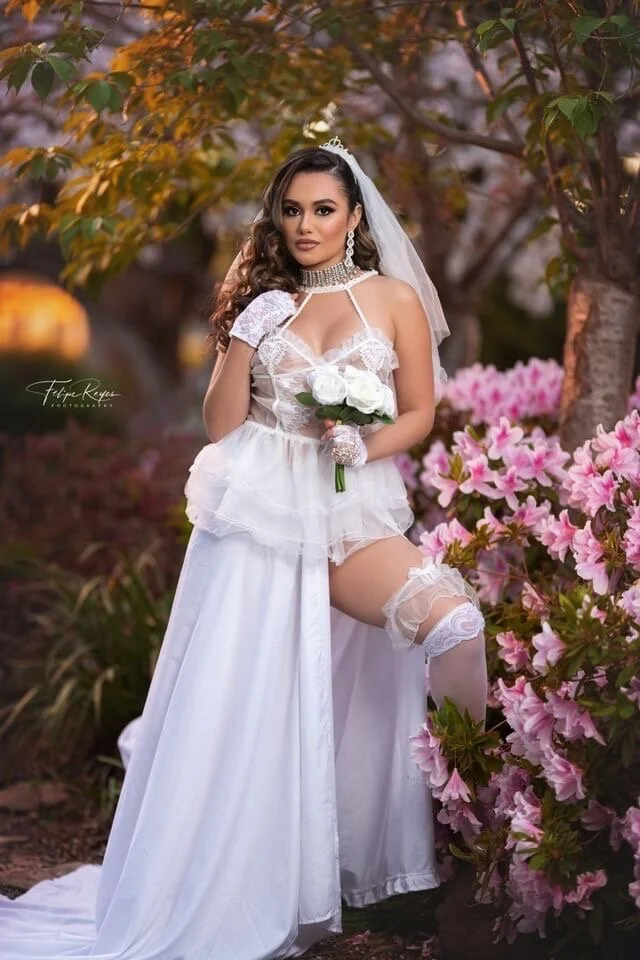 A woman dressed in a wedding gown and veil, holding a bouquet of white roses, standing outdoors among pink azalea bushes and trees with autumn-colored leaves.