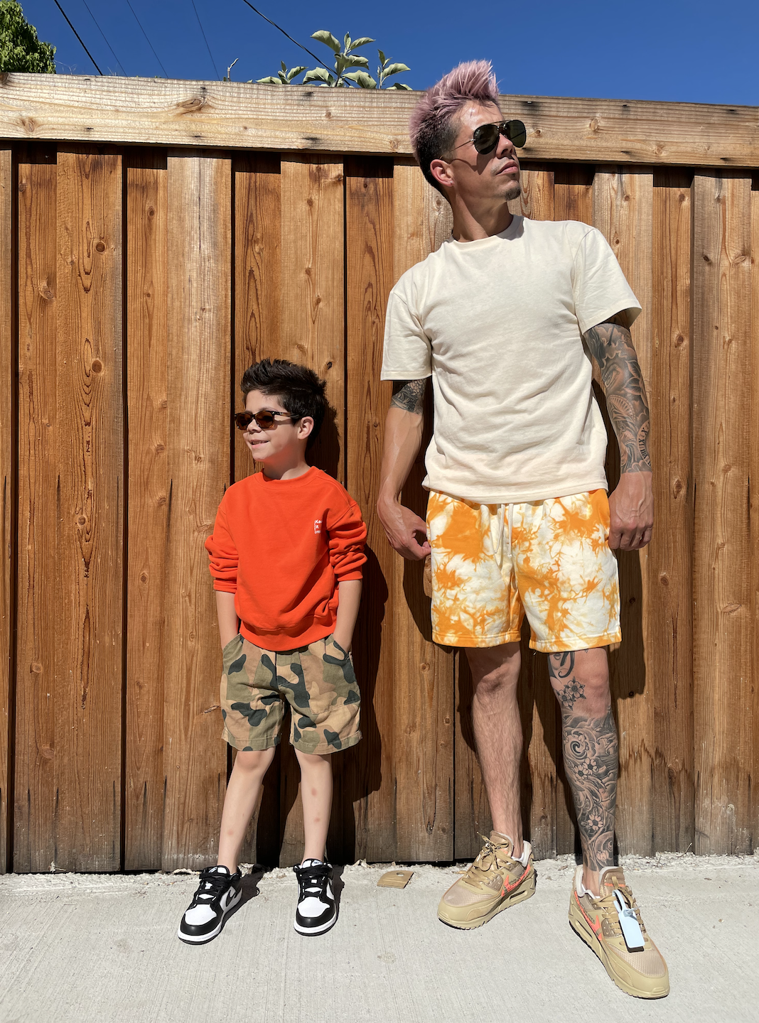 A man and a boy standing in front of a wooden fence and looking to the side. The man wears sunglasses, a beige t-shirt, yellow tie-dye shorts, and sneakers, and has tattoos on his arms and leg. The boy wears sunglasses, a red sweatshirt, camouflage s