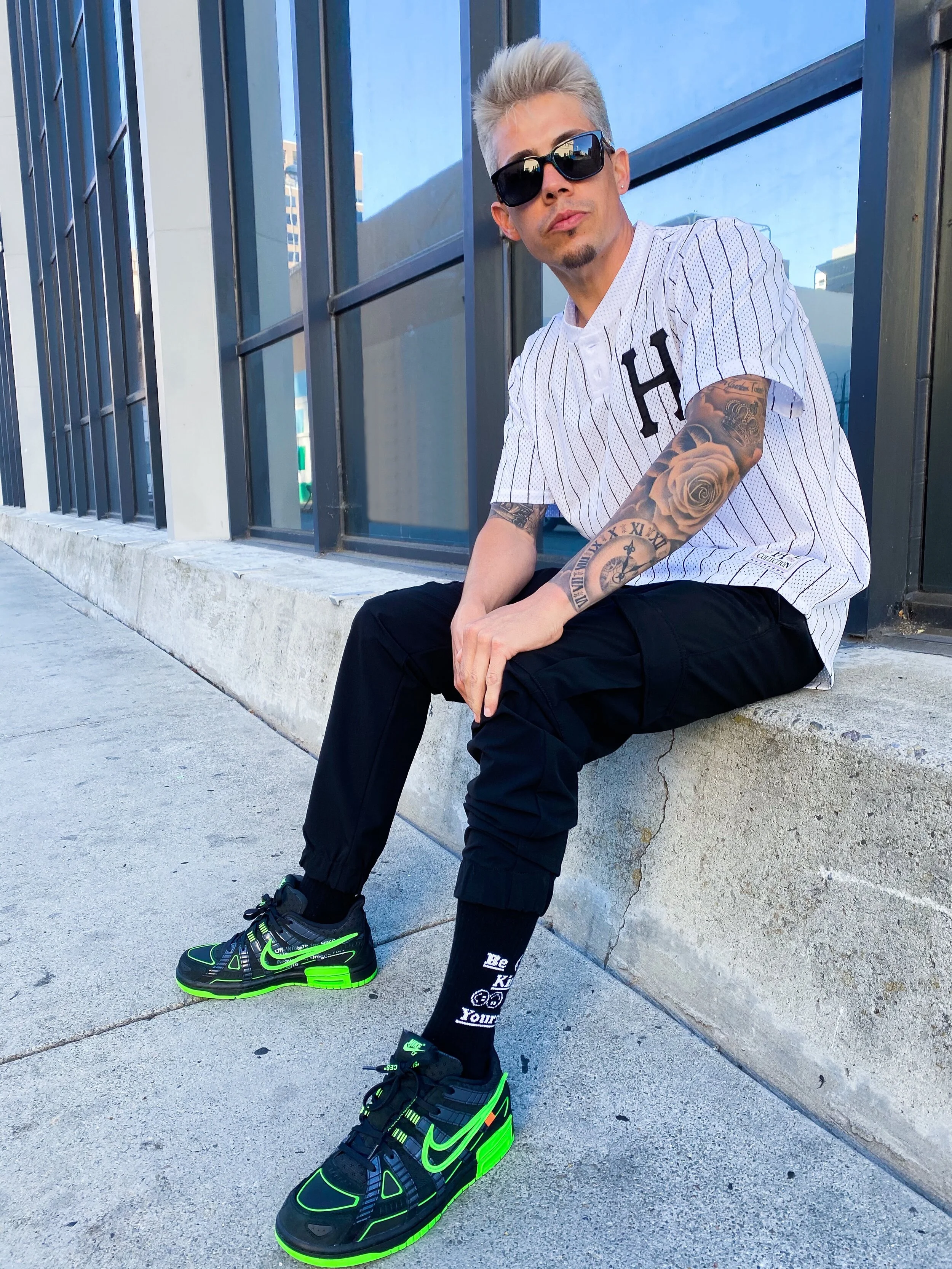 A young man with platinum blond hair, wearing sunglasses, a white pinstripe jersey with a large letter 'H' on it, black pants, and neon green and black Nike sneakers, sitting on a concrete ledge outside a modern building with large glass windows.